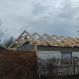 Konstrukcja drewniana dachu w trakcie budowy na przyczepie kempingowej, widok z dołu na tle pochmurnego nieba i zarośli.