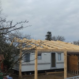 Mar-Kuc - Konstrukcja drewniana dachu w trakcie budowy nad domkiem holenderskim, widoczna drabina, drzewa i budynki w tle, pochmurne niebo.