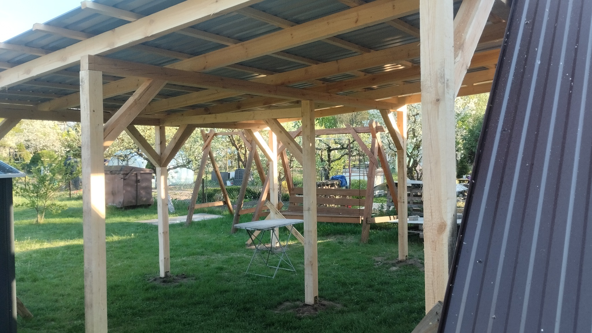 Konstrukcja drewnianej altany z metalowym dachem w ogrodzie, widok na huśtawkę i stół. Część blachy dachowej widoczna z boku.
