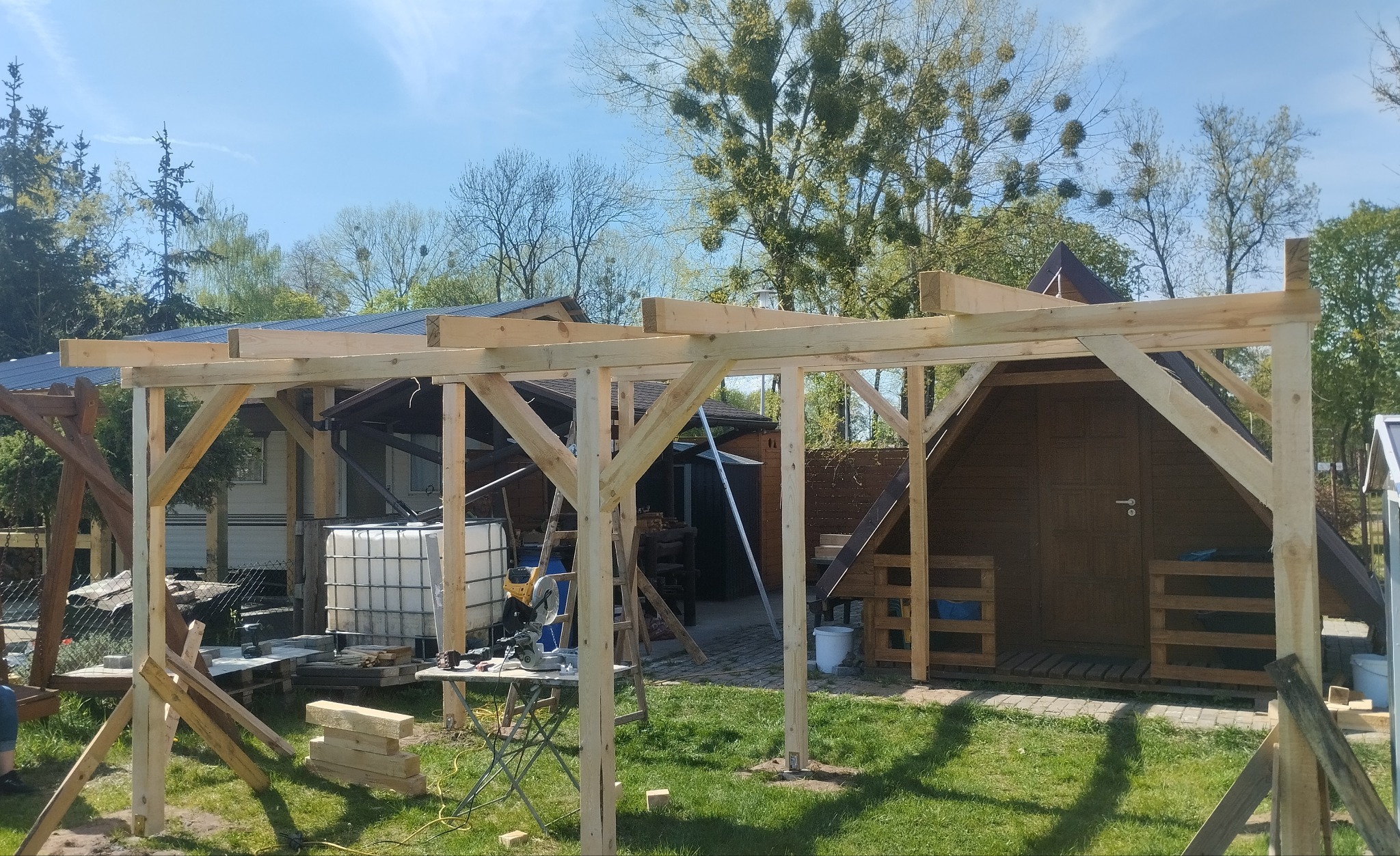 Drewniana konstrukcja z jasnego drewna w ogrodzie, budowa altany w toku. W tle domki, drzewa i niebieskie niebo.