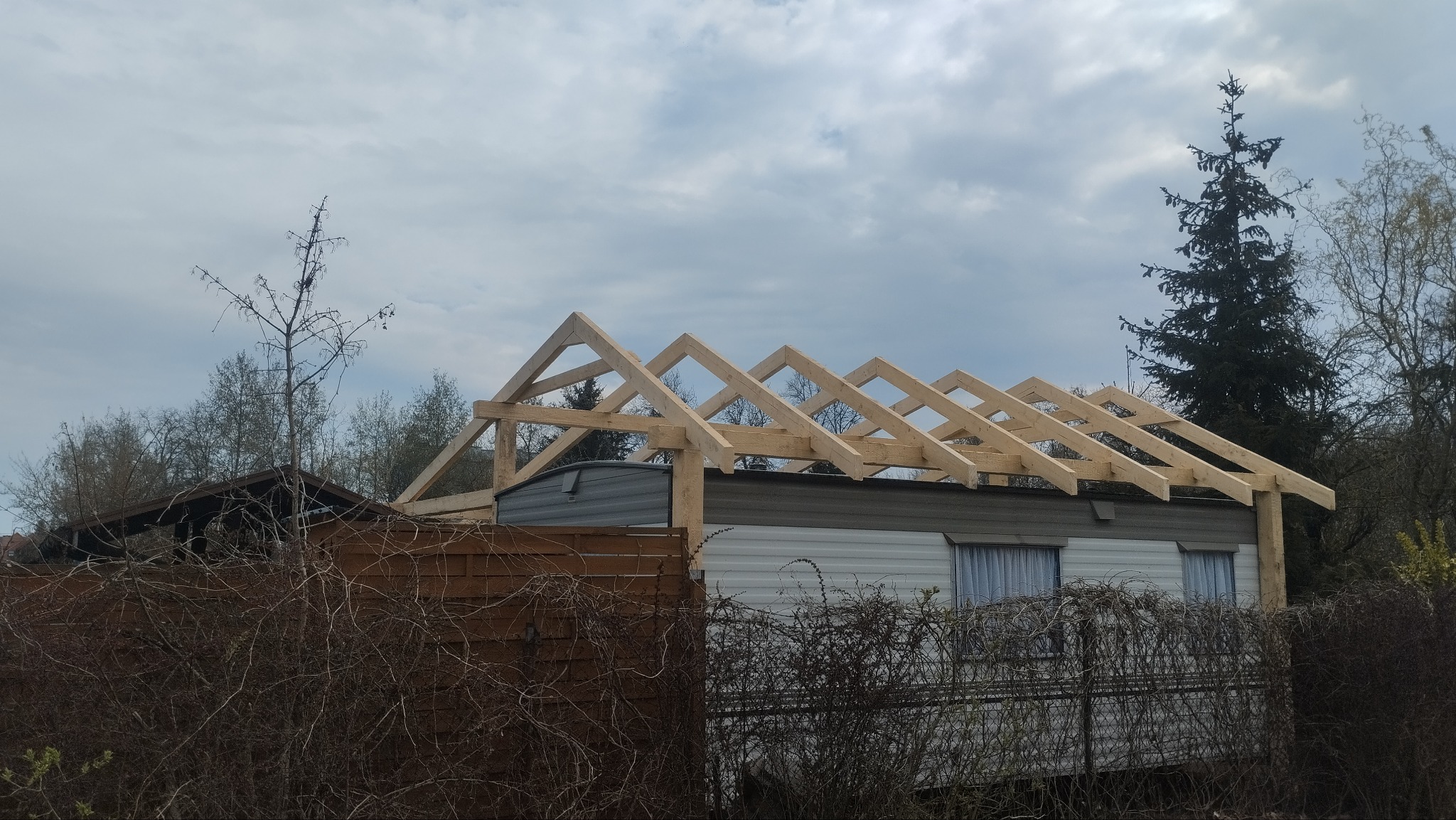 Konstrukcja drewniana dachu w trakcie budowy na przyczepie kempingowej, widok z dołu na tle pochmurnego nieba i zarośli.