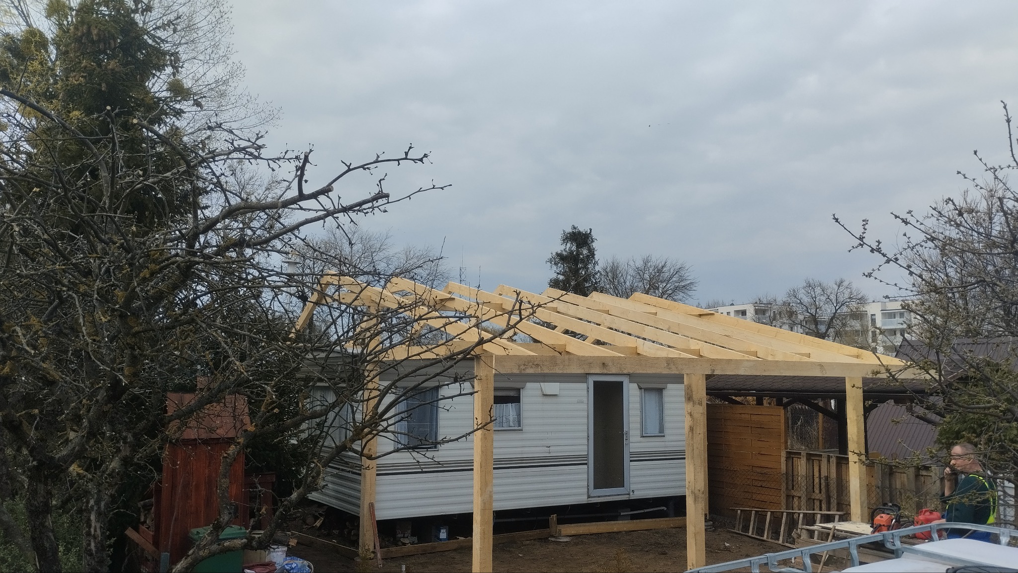 Konstrukcja drewniana dachu w trakcie budowy nad domkiem holenderskim, widoczna drabina, drzewa i budynki w tle, pochmurne niebo.