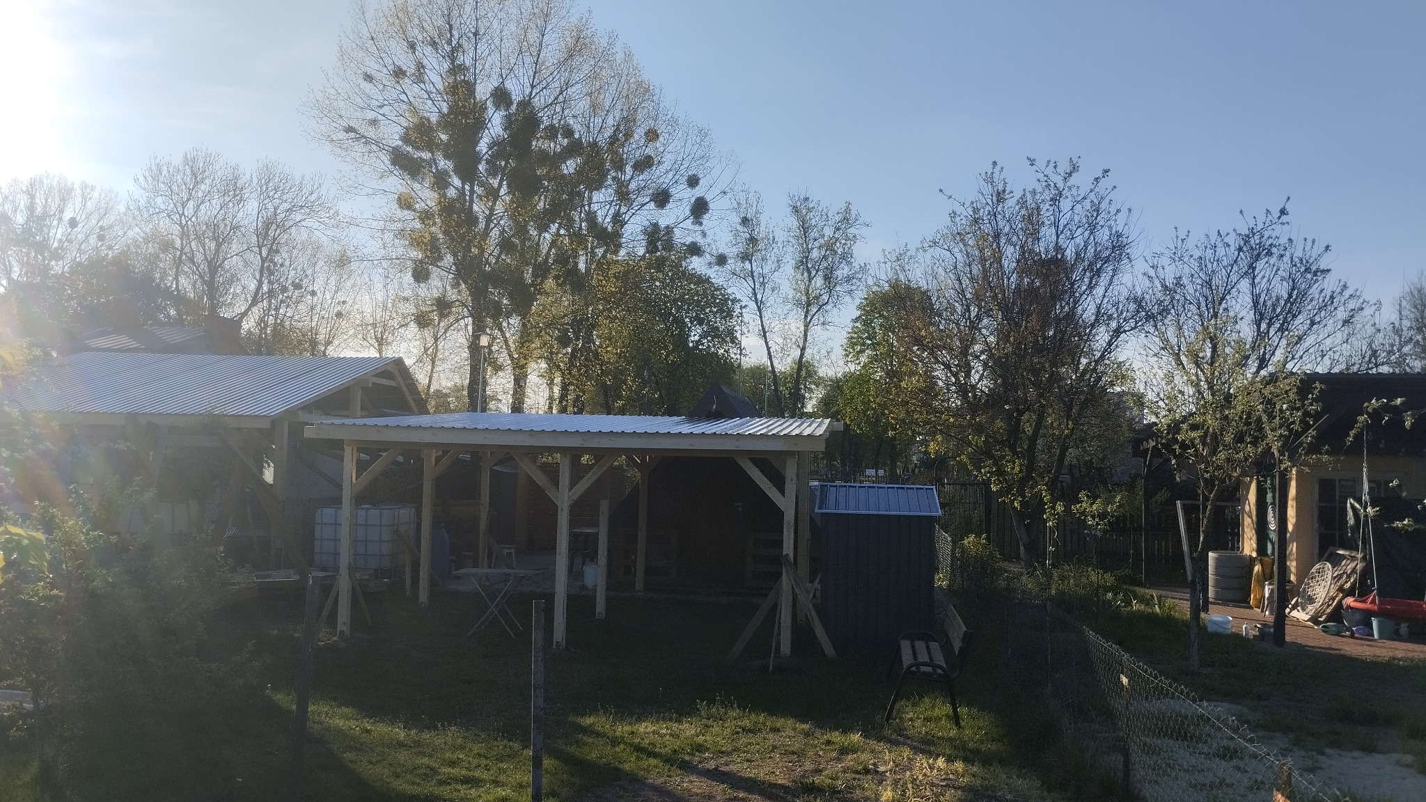 Zadaszenie z blachy trapezowej wsparte na drewnianej konstrukcji, otoczone zielenią ogrodu, w tle drzewa i budynki. Widok w słoneczny dzień.