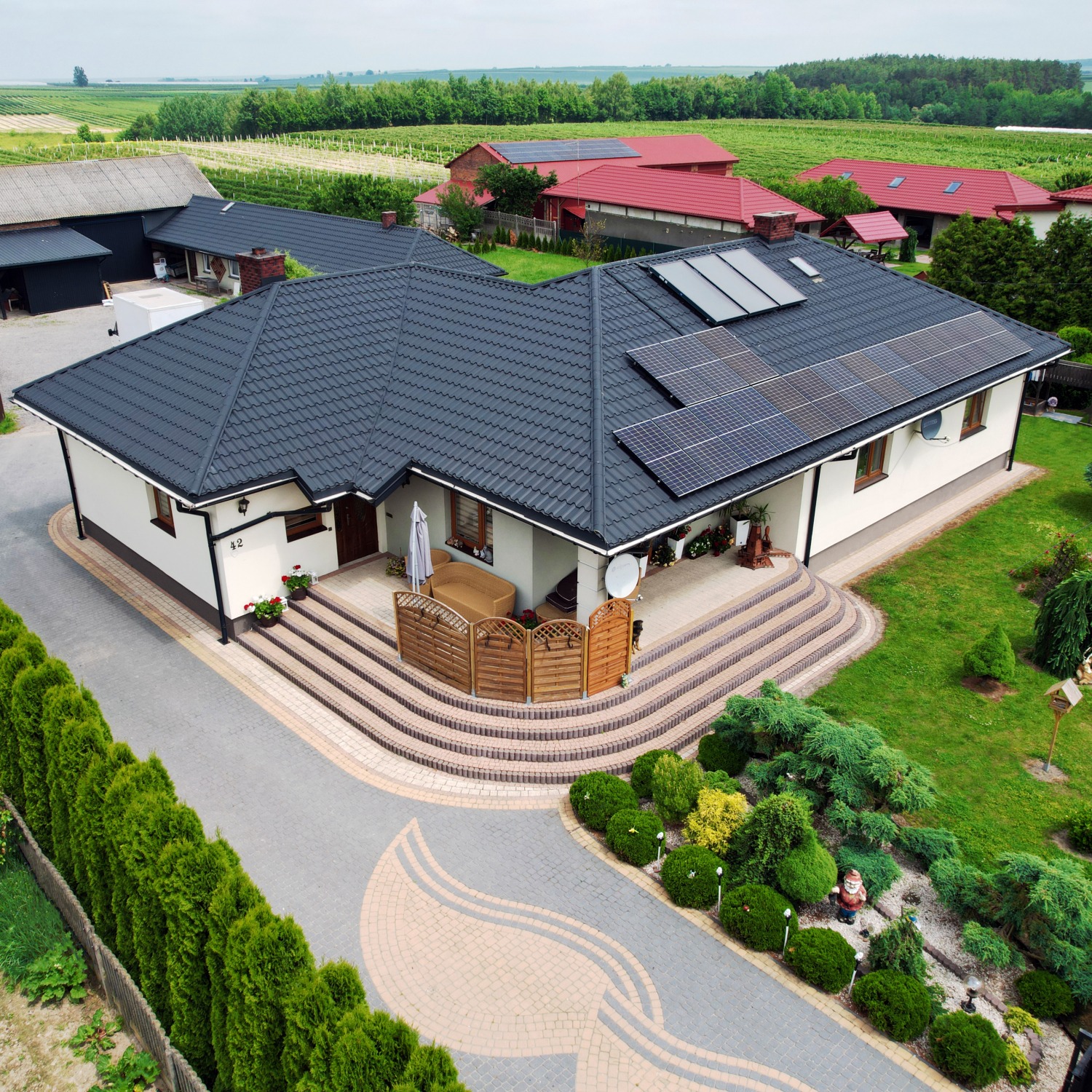 Dom jednorodzinny z ciemną dachówką i panelami fotowoltaicznymi na dachu, widok z góry, zadbany ogród z tujami i ozdobnymi krzewami, kostka brukowa na podjeździe.