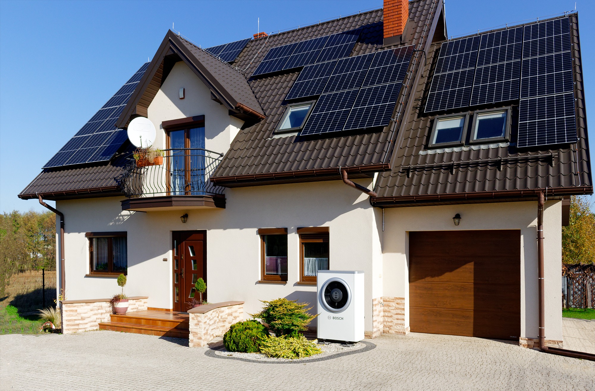 Dom jednorodzinny z brązowym dachem, na którym zamontowane są panele słoneczne; na trawniku stoi biała pompa ciepła marki Bosch.