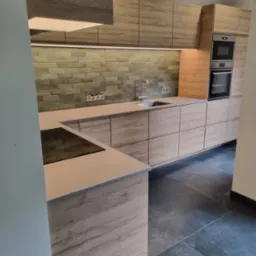 Nowoczesna kuchnia z drewnianymi frontami, szarymi płytkami na ścianie i ciemną podłogą z dużych płytek. Widoczny zlew ze stali nierdzewnej, płyta indukcyjna i wbudowany piekarnik z mikrofalówką.