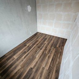 Dariusz Zambrzycki DMM Usługi Glazurnicze - Ułożona podłoga z paneli imitujących ciemne drewno w małym pomieszczeniu w trakcie remontu, ściany częściowo otynkowane, widoczna surowa żarówka.