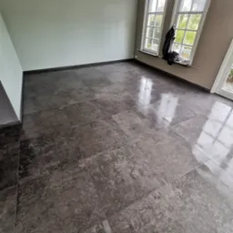 Ułożone, ciemnoszare, matowe płytki podłogowe w pustym pokoju z dwoma oknami i jasnymi ścianami.