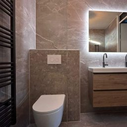 Dariusz Zambrzycki DMM Usługi Glazurnicze - Nowoczesna łazienka z marmurowymi płytkami, podwieszaną toaletą i podświetlanym lustrem. Minimalistyczny design, brązowe odcienie, grzejnik drabinkowy.