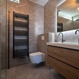 Dariusz Zambrzycki DMM Usługi Glazurnicze - Elegancka łazienka z szarymi płytkami imitującymi marmur, podwieszana toaleta, drewniana szafka z umywalkami i czarny grzejnik drabinkowy. Oświetlenie LED.