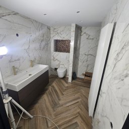 Dariusz Zambrzycki DMM Usługi Glazurnicze - Wykończona łazienka z marmurowymi płytkami, drewnianą podłogą w jodełkę i nowoczesną umywalką. Widoczne otwory na oświetlenie w suficie i dekoracyjna mozaika.