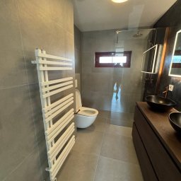 Dariusz Zambrzycki DMM Usługi Glazurnicze - Nowoczesna łazienka w szarościach z białym grzejnikiem drabinkowym, umywalkami nablatowymi i kabiną prysznicową typu walk-in. Widoczne płytki ceramiczne na ścianach i podłodze.