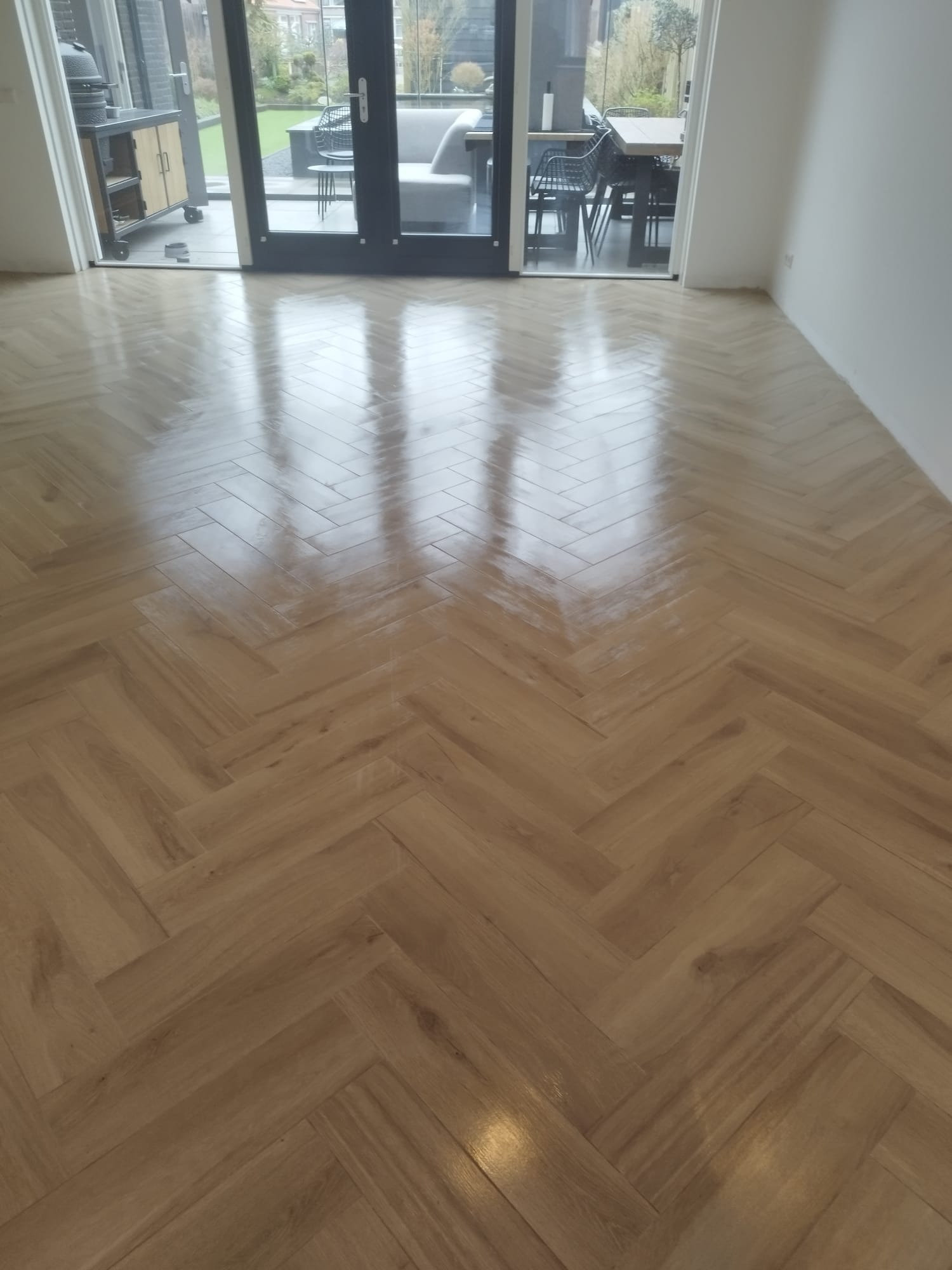 Ułożona w jodełkę, błyszcząca podłoga z paneli imitujących drewno, z refleksami światła z okna tarasowego, widok na zewnętrzny taras z meblami ogrodowymi.