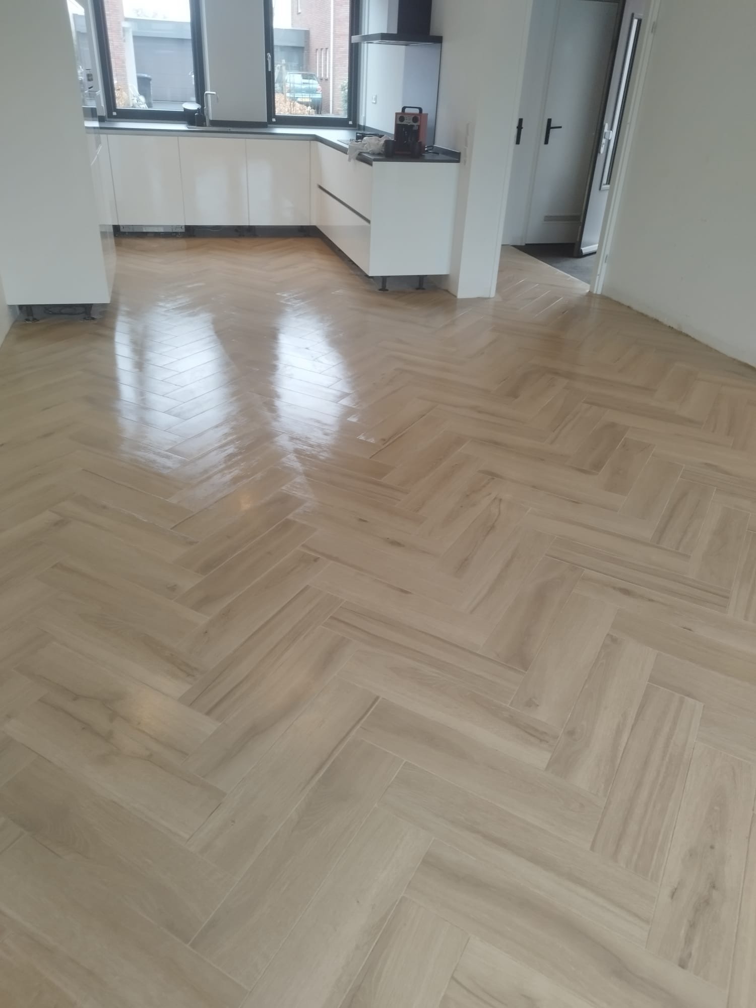 Jasna, błyszcząca podłoga ułożona w jodełkę w nowym, minimalistycznym wnętrzu kuchni i przedpokoju. Widoczne białe szafki kuchenne bez uchwytów i fragment drzwi.