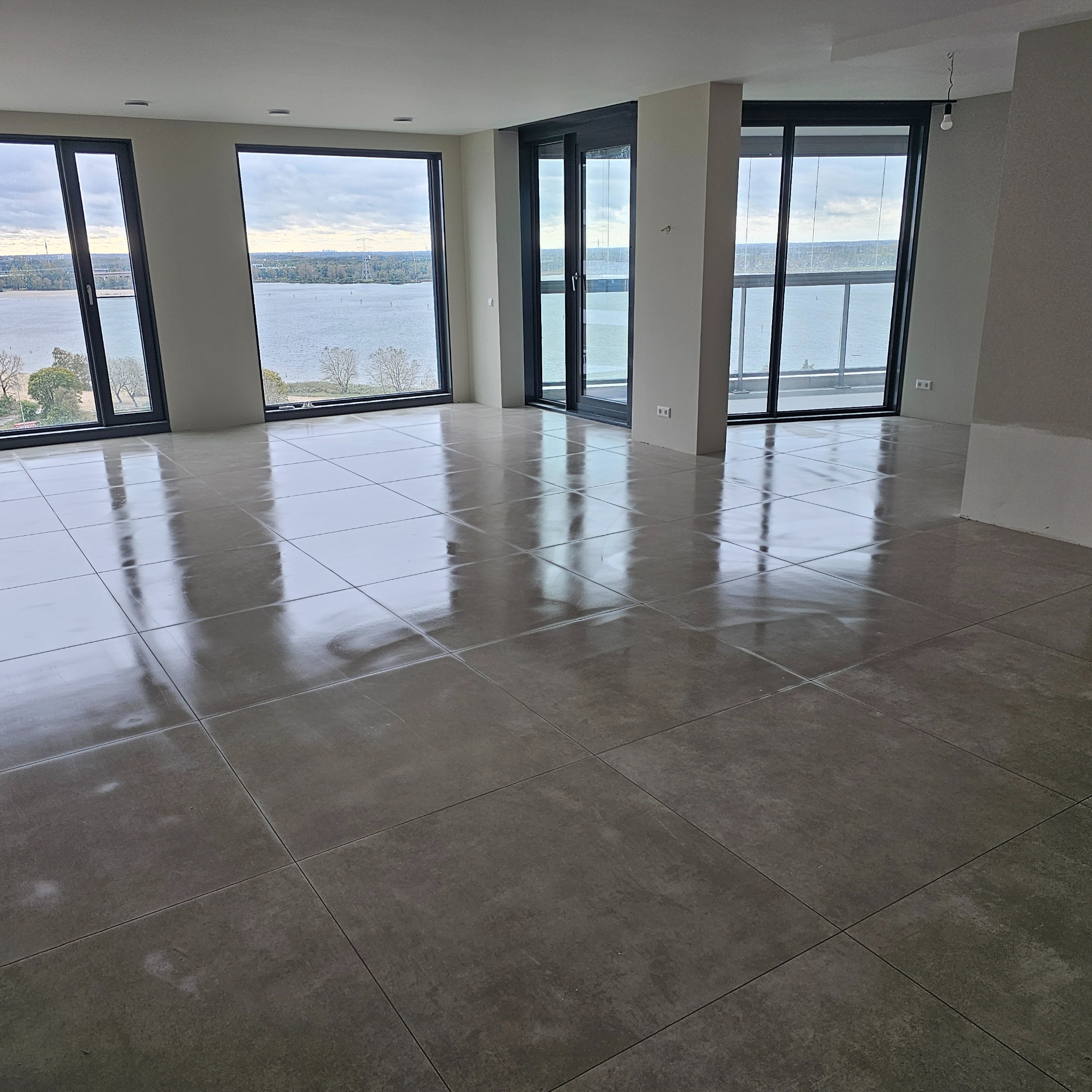 Wykończone wnętrze z dużymi, szarymi płytkami podłogowymi i panoramicznymi oknami z widokiem na rzekę.
