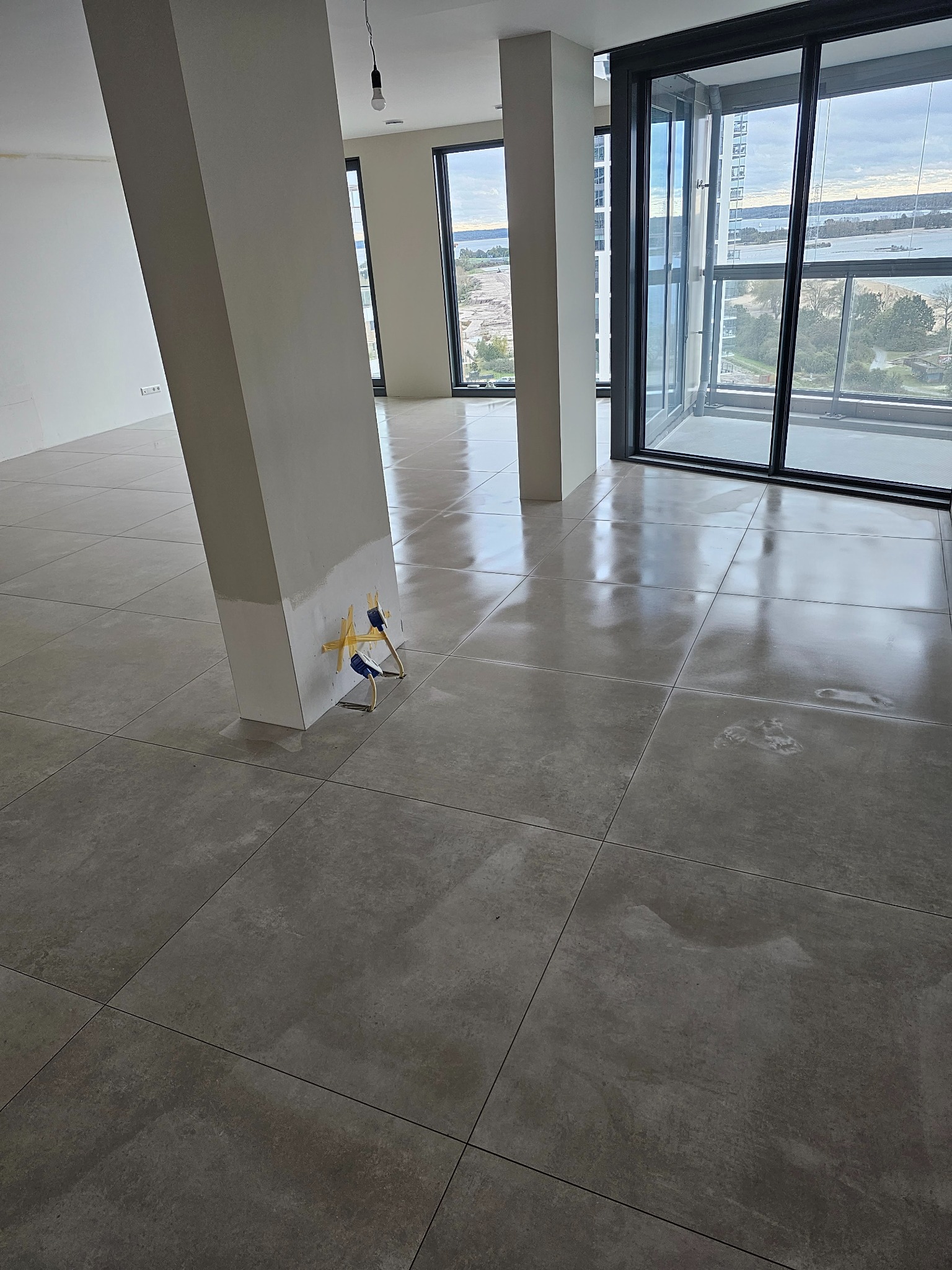 Nowo ułożone, duże, szare płytki podłogowe w przestronnym wnętrzu z widokiem na miasto przez duże okna; widoczne kliny poziomujące przy słupie konstrukcyjnym.