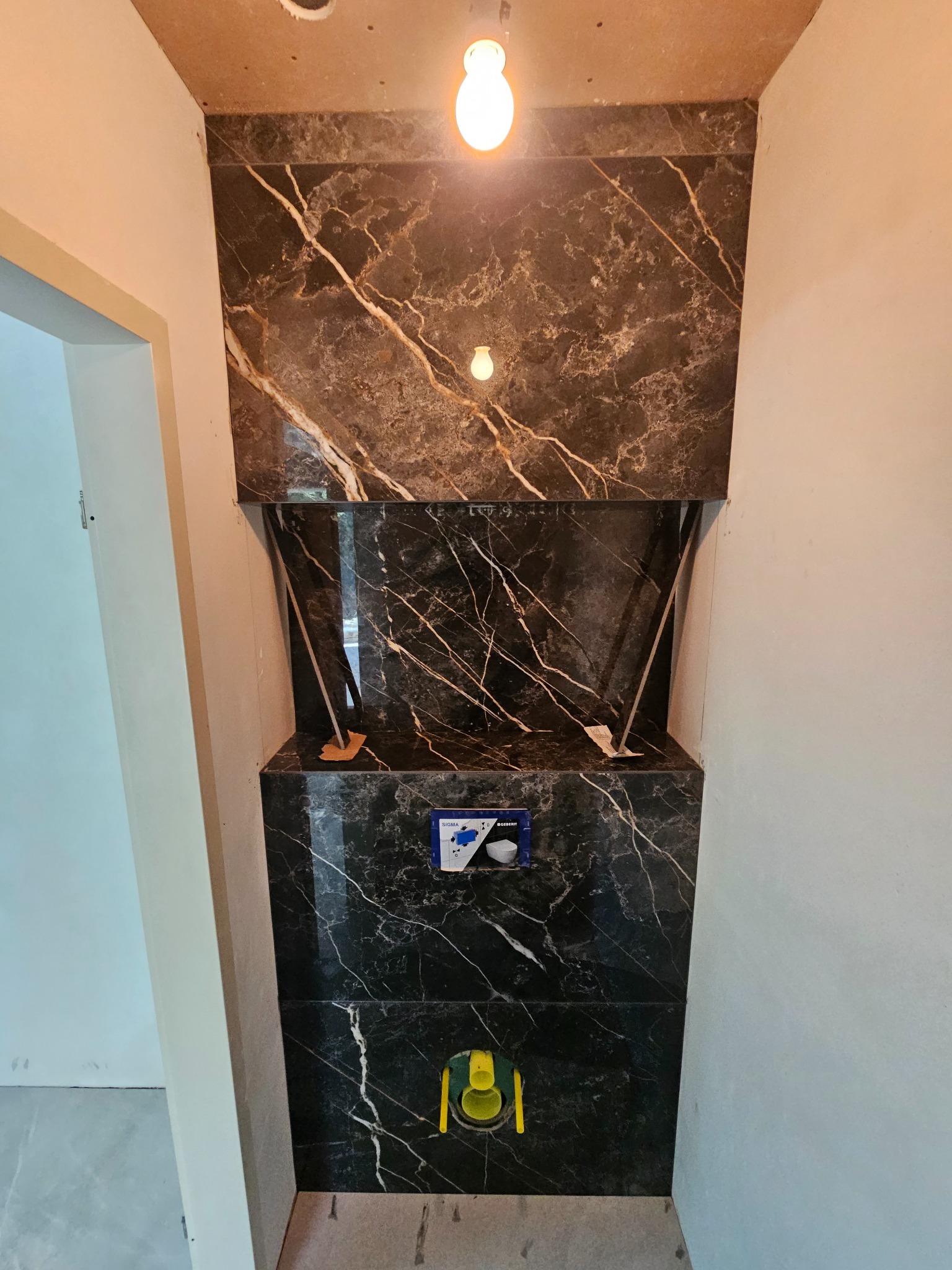 Wykończona wnęka w łazience z czarnymi płytkami imitującymi marmur z białymi żyłami; widoczny stelaż podtynkowy WC z przyciskiem Geberit i żółta zaślepka kanalizacyjna.