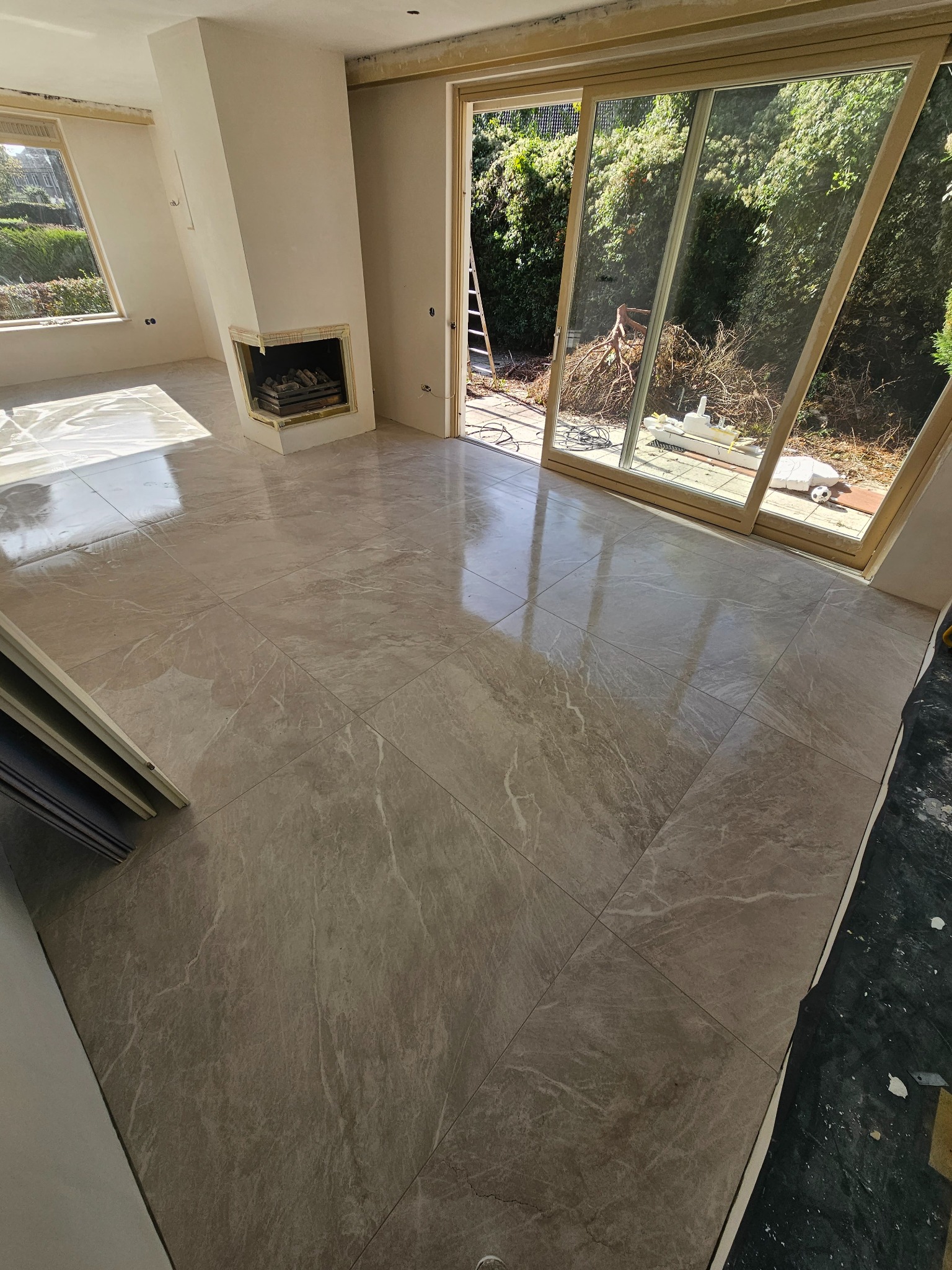 Beżowe płytki podłogowe imitujące marmur w jasnym pokoju z kominkiem i dużym oknem tarasowym, widoczne ślady prac wykończeniowych.