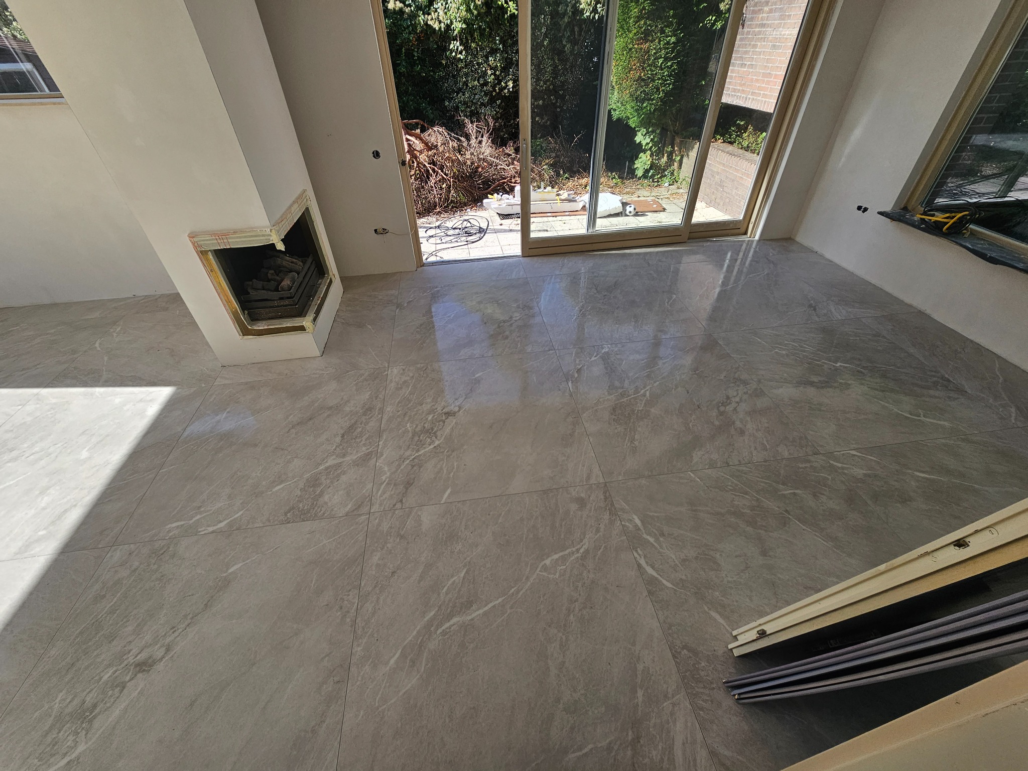 Ułożone szare płytki imitujące marmur na podłodze w salonie z kominkiem, widoczne otwarte drzwi balkonowe i częściowo zakryte listwy przypodłogowe.