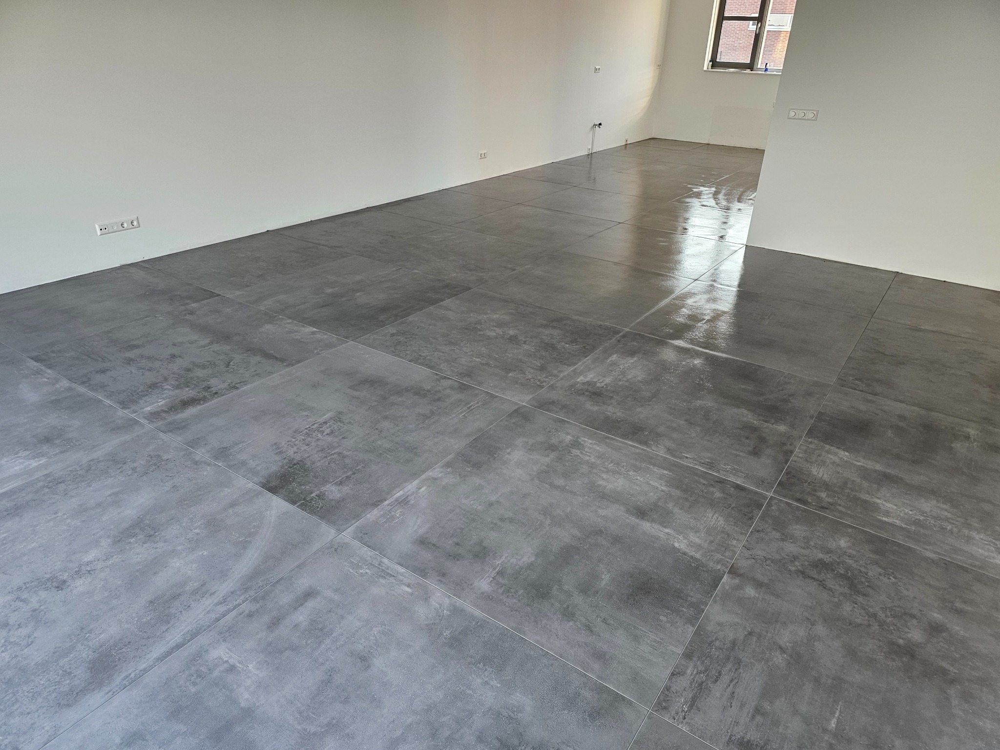Świeżo położone, duże, szare płytki gresowe w pustym pomieszczeniu w Opolu, jeszcze mokre po myciu, odbijające światło z okna.