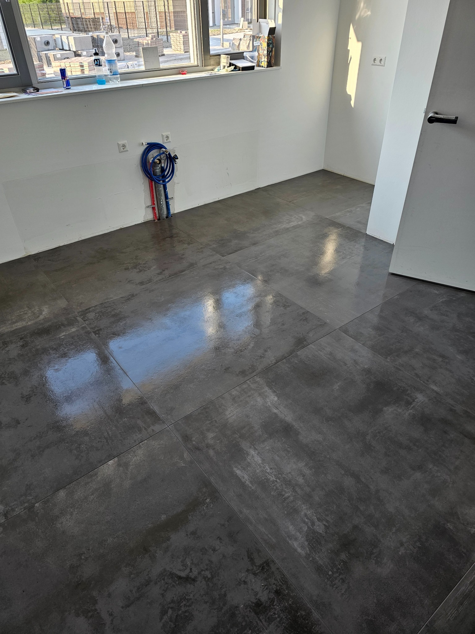 Ułożona, ciemnoszara, błyszcząca podłoga z płytek w surowym wnętrzu łazienki w trakcie remontu, widoczne wyprowadzenia instalacji wodnej na ścianie.