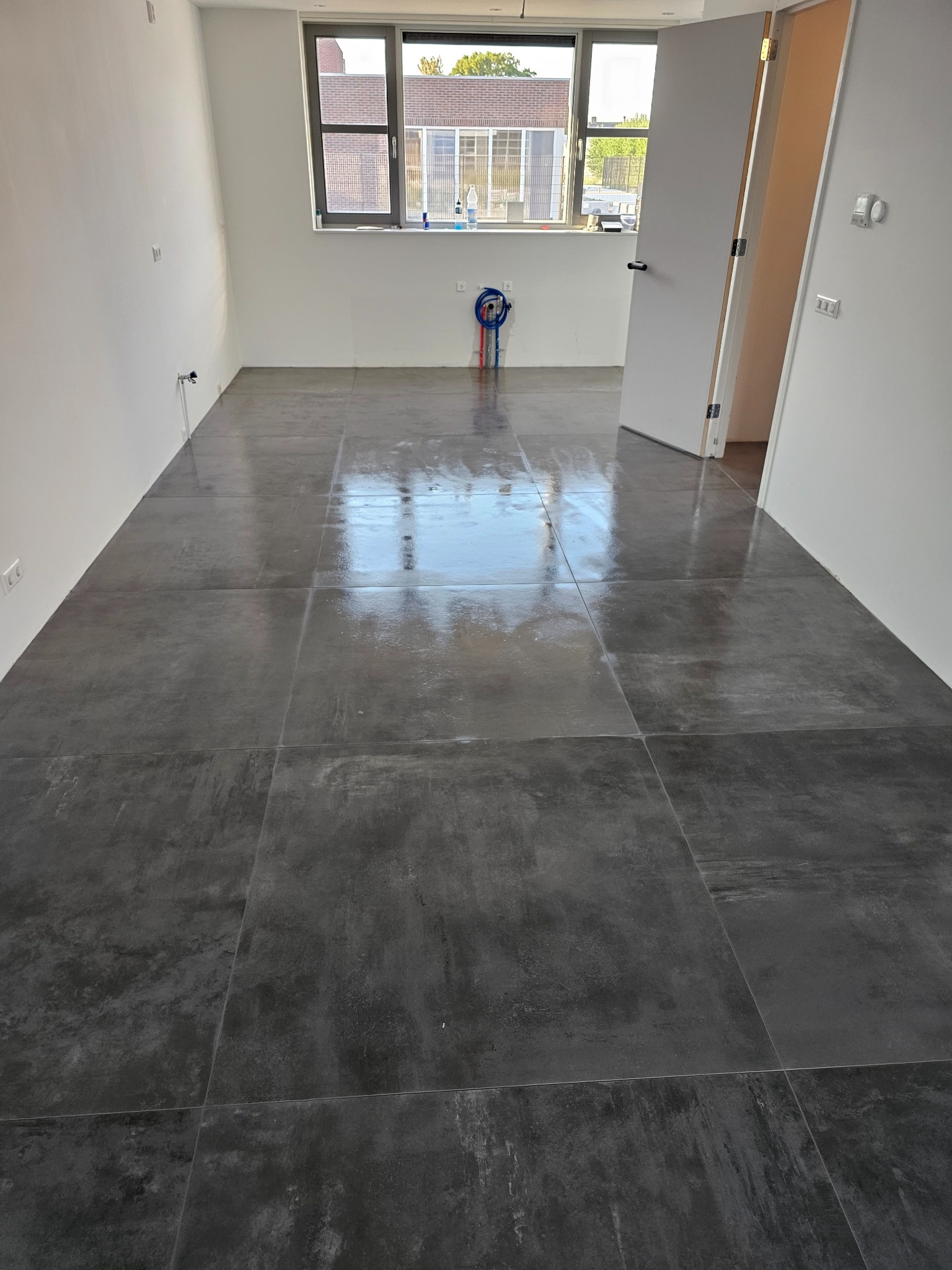 Ułożone, ciemnoszare, błyszczące płytki podłogowe w pustym pomieszczeniu z białymi ścianami i otwartymi białymi drzwiami. Widoczne instalacje wodne na ścianie pod oknem.