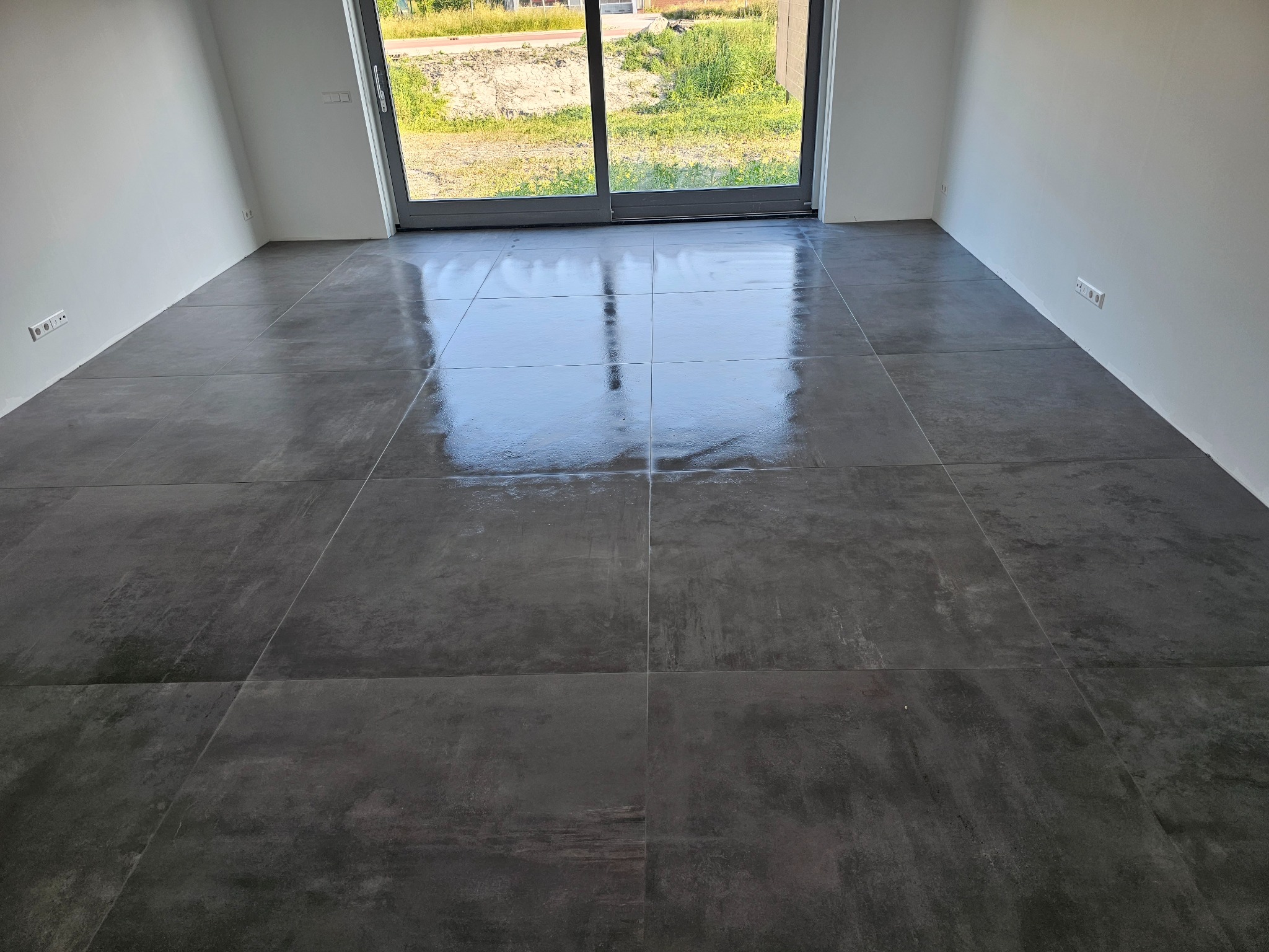 Ułożone, ciemnoszare płytki podłogowe z efektem betonu w pustym pomieszczeniu z dużym oknem balkonowym, widoczne gniazdka elektryczne przy ścianie.
