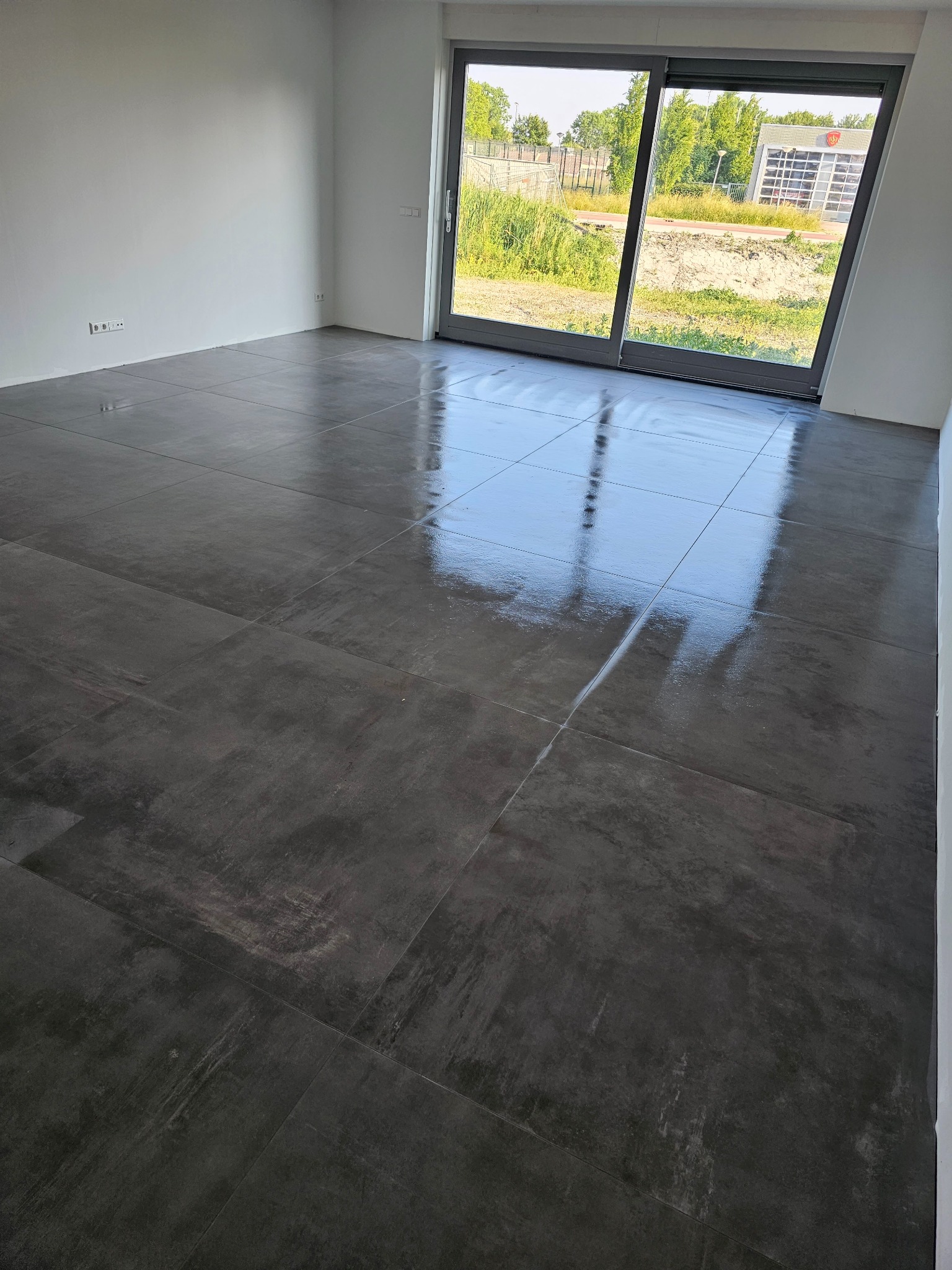 Świeżo położona, ciemnoszara, błyszcząca podłoga z płytek ceramicznych w pustym pomieszczeniu z dużym przesuwnym oknem, odbijającym budynek w tle.