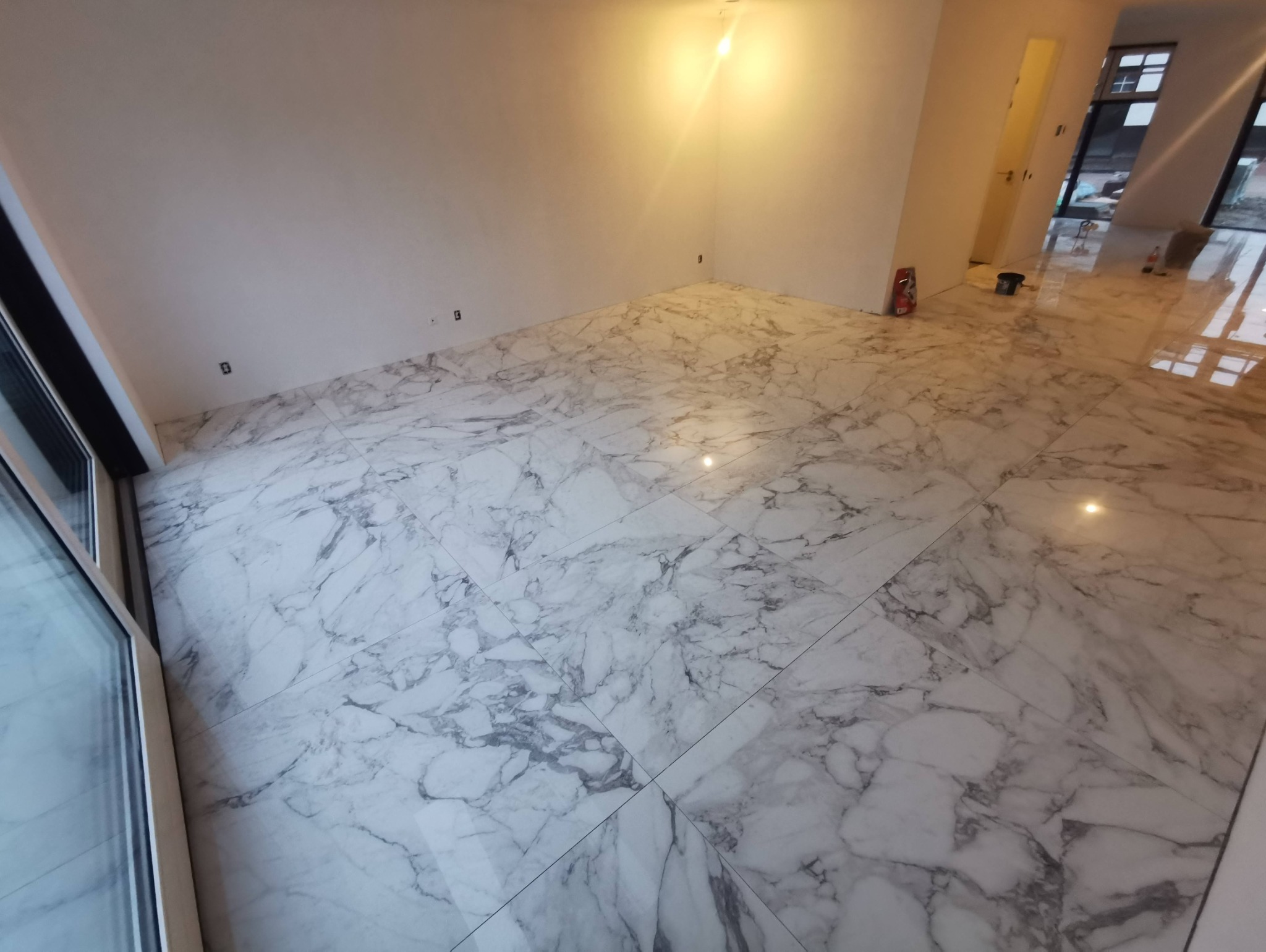 Wykończona podłoga z płytek imitujących marmur Carrara w jasnym, pustym pomieszczeniu z dużym oknem i widocznymi narzędziami glazurniczymi w tle.