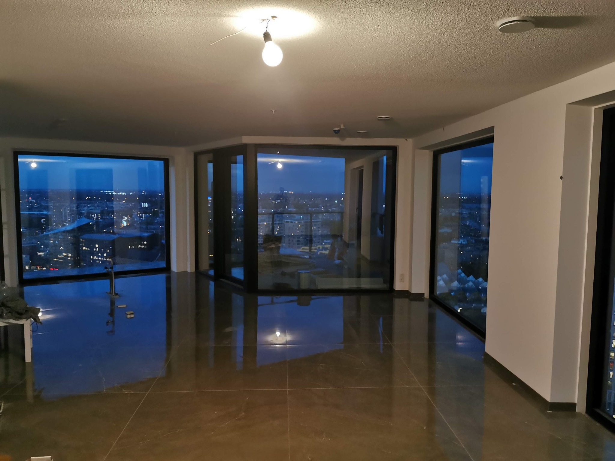 Przestronne, puste wnętrze z dużymi oknami z widokiem na nocne miasto, świeżo położona, błyszcząca podłoga z ciemnych płytek imitujących kamień, surowe oświetlenie żarówką.