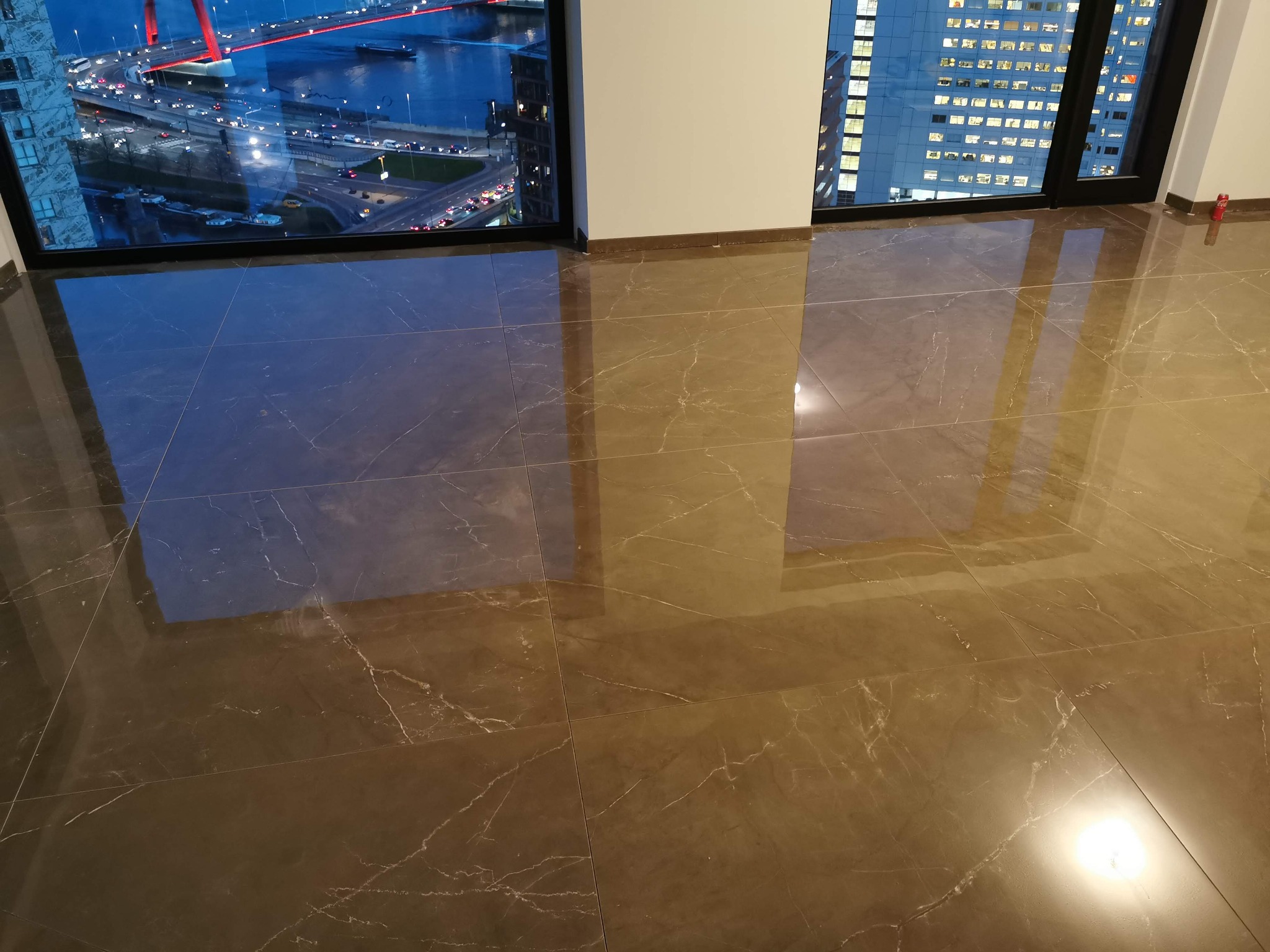 Błyszcząca, brązowa podłoga z płytek imitujących marmur, z widocznym odbiciem nocnego krajobrazu miejskiego za oknem.