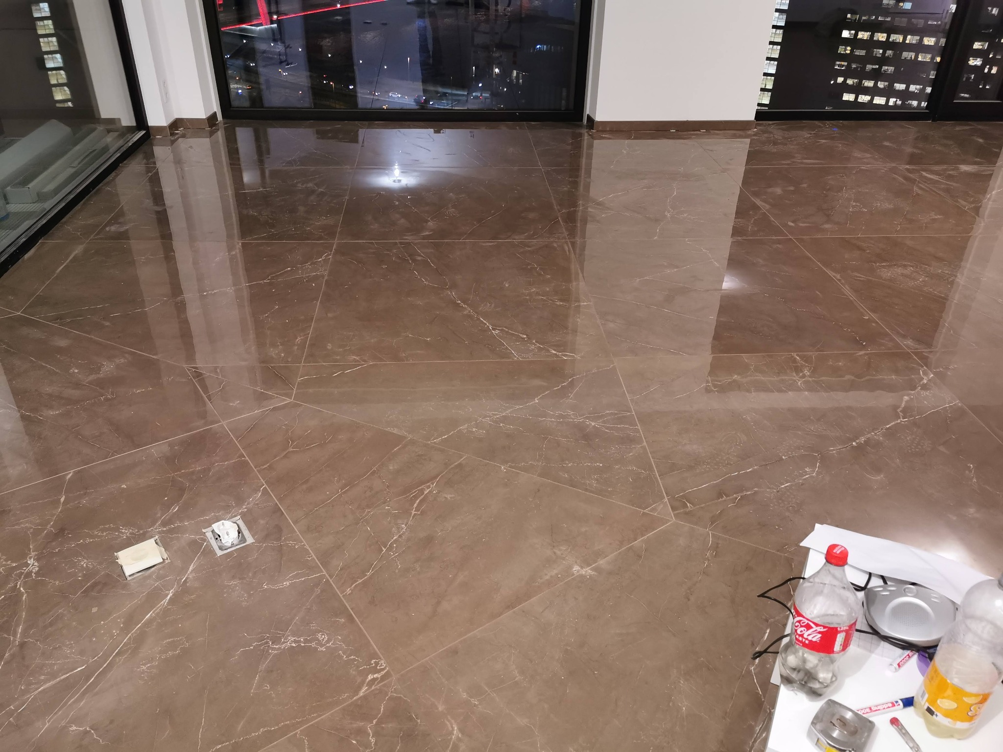 Błyszcząca, brązowa podłoga z płytek imitujących marmur, widoczne dwa otwory instalacyjne bez osprzętu, butelka Coca-Coli i inne akcesoria na kartce papieru w rogu pokoju.