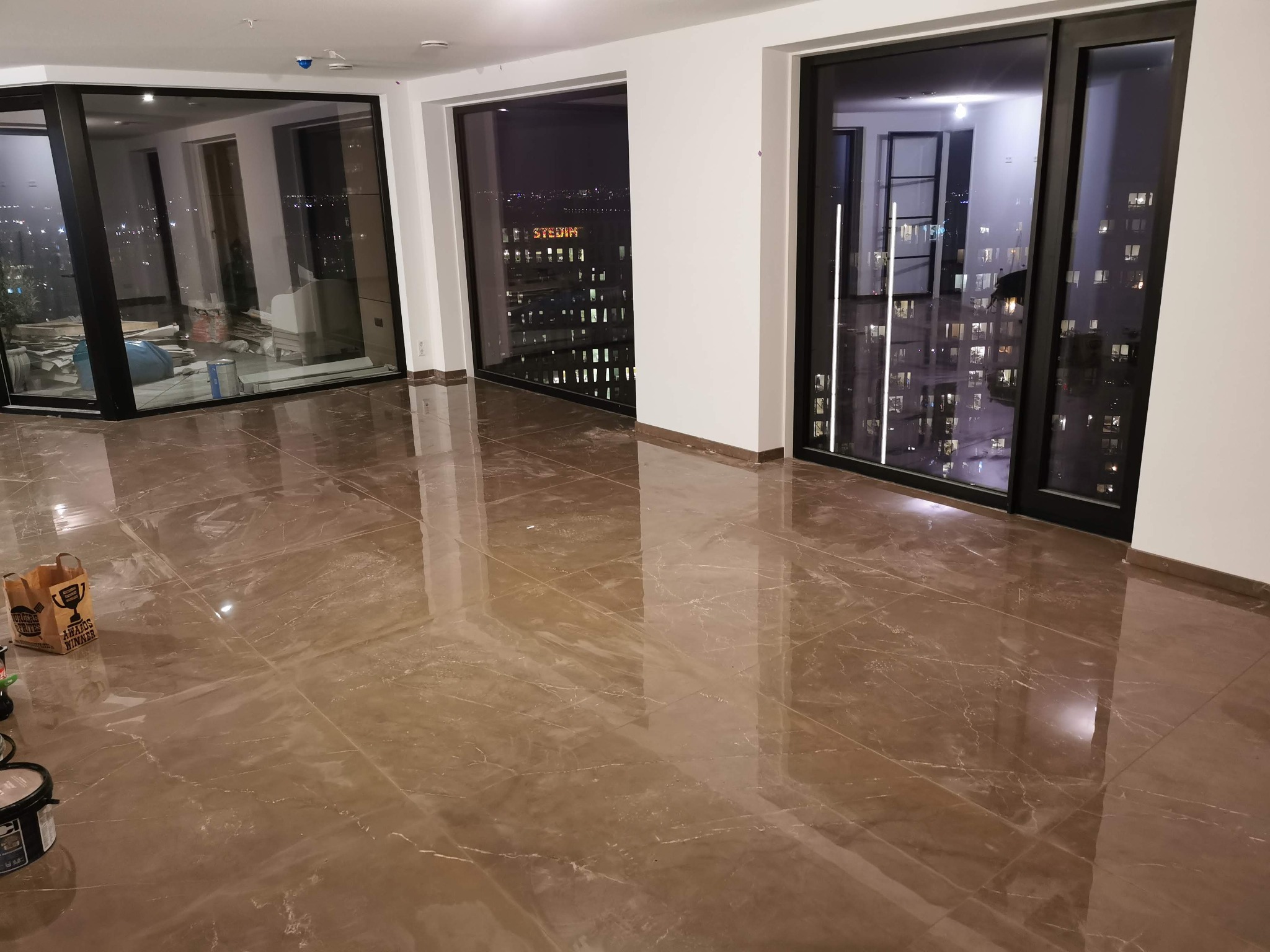 Błyszcząca, brązowa podłoga z płytek imitujących marmur w nowym apartamencie z dużymi oknami z widokiem na nocne miasto. Widoczne wiaderko z farbą i torba z zakupami budowlanymi w rogu pokoju.