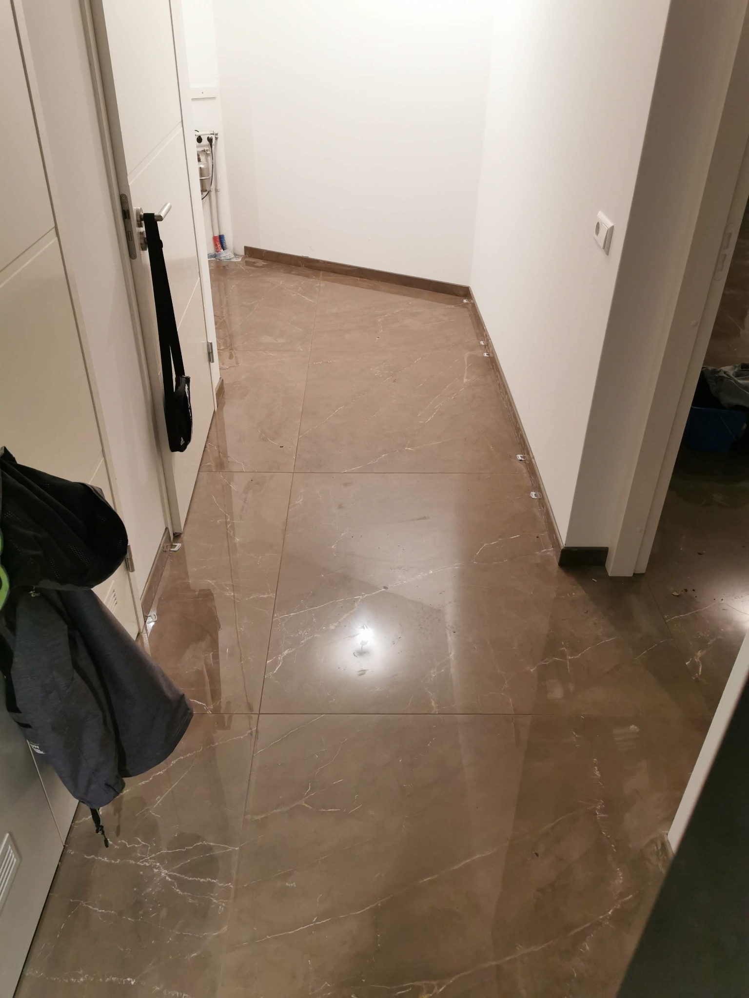 Błyszcząca, brązowa podłoga z płytek imitujących marmur w korytarzu, widoczne białe ściany i drzwi, plastikowe krzyżyki do fugowania pomiędzy płytkami przy ścianie.
