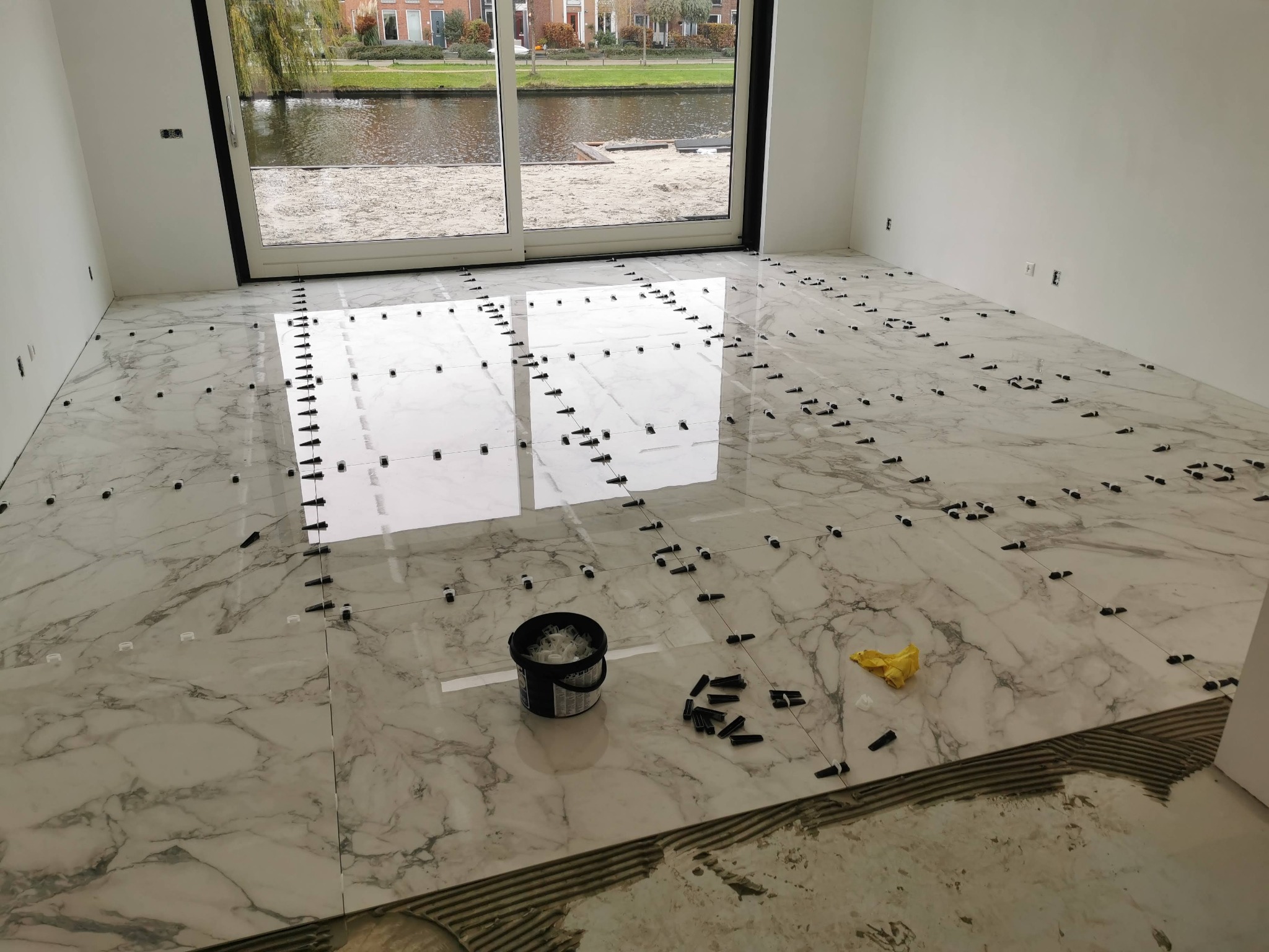 Układanie płytek marmuropodobnych w jasnym pomieszczeniu z widokiem na kanał przez duże okno; widoczne czarne kliny i system poziomowania płytek, wiadro z elementami montażowymi oraz ślady kleju...