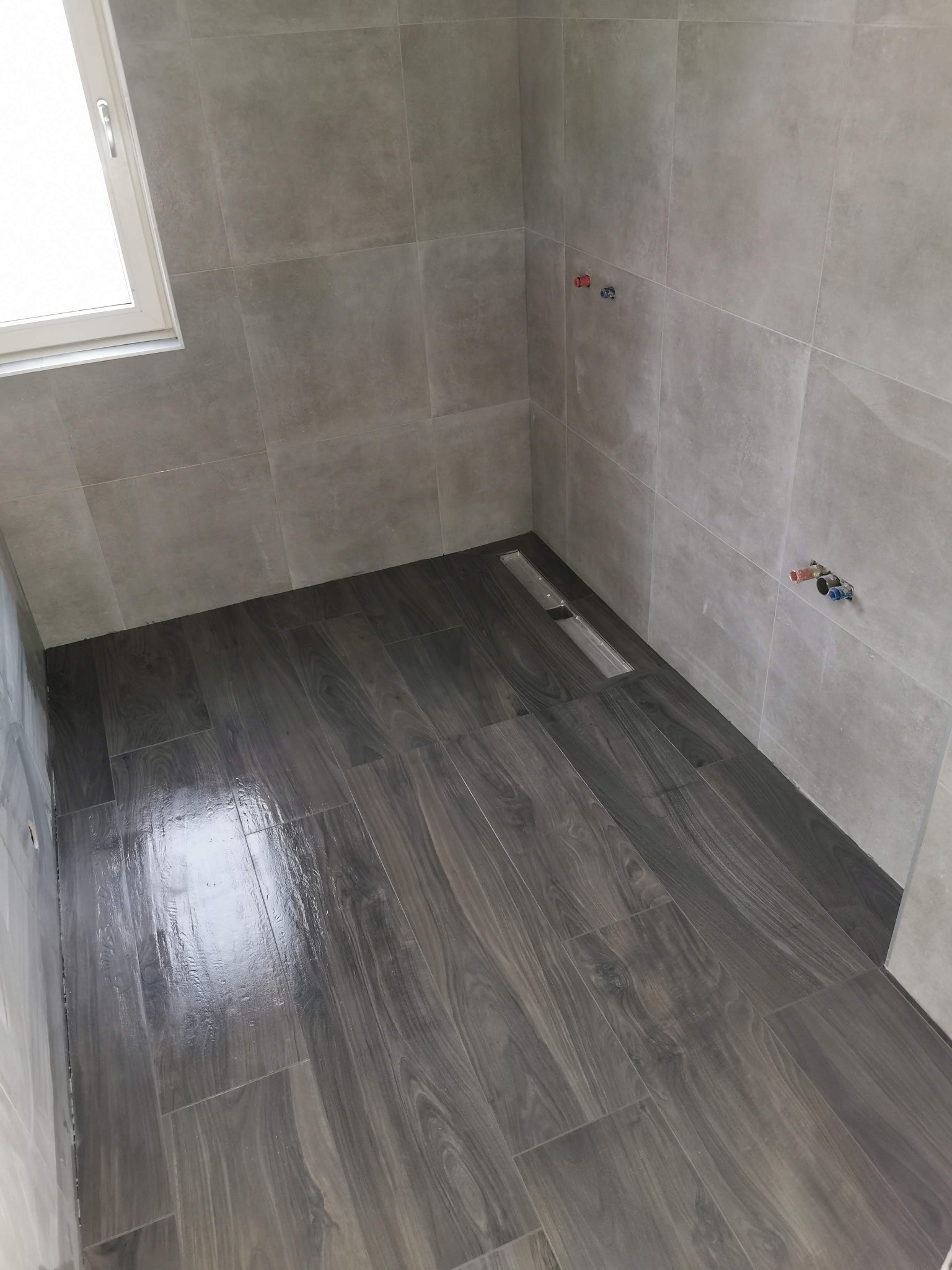 Nowoczesna kabina prysznicowa wykończona szarymi płytkami ceramicznymi ściennymi i podłogowymi imitującymi drewno, z odpływem liniowym w podłodze i widocznymi przyłączami wodnymi na ścianie.