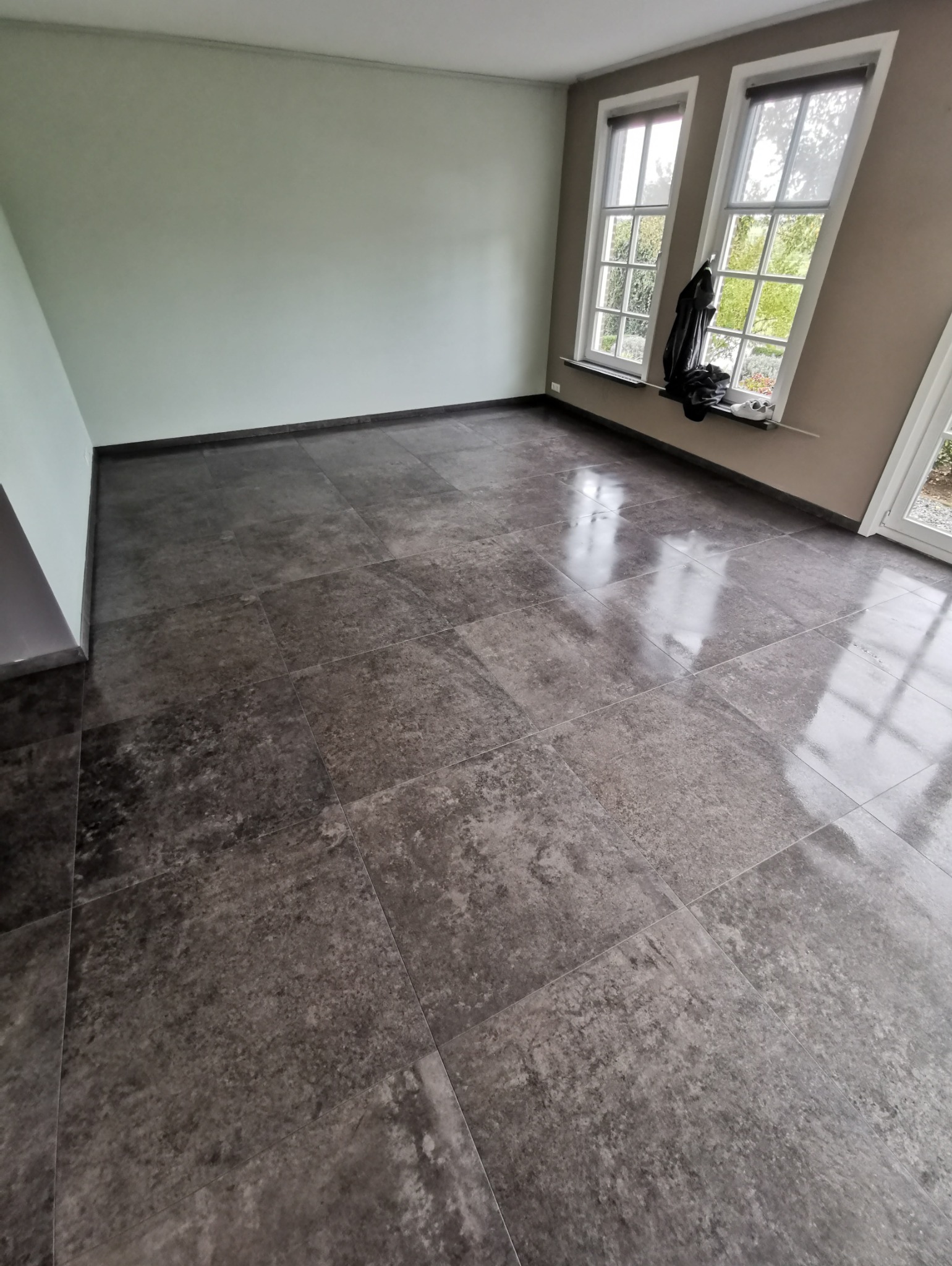 Ułożone, ciemnoszare, matowe płytki podłogowe w pustym pokoju z dwoma oknami i jasnymi ścianami.