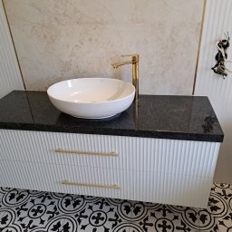 SZPAK MEBLE - Łazienkowa szafka pod umywalkę z czarnym blatem i białymi, ryflowanymi frontami szuflad, złota bateria i umywalka nablatowa w Radomsku, na podłodze wzorzyste płytki.