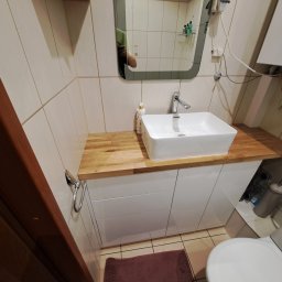 SZPAK MEBLE - Nowoczesna łazienka z białą, prostokątną umywalką nablatową na drewnianym blacie i białymi szafkami, widoczne odbicie osoby w lustrze, brązowy dywanik na podłodze.