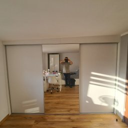 SZPAK MEBLE - Szafa wnękowa z przesuwnymi, białymi drzwiami z lustrem, w którym odbija się osoba robiąca zdjęcie oraz biurko z krzesłem w pokoju.