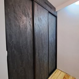 SZPAK MEBLE - Nowoczesna szafa przesuwna z ciemnoszarymi drzwiami o teksturze kamienia, drewniana podłoga w jasnym odcieniu.