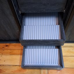 SZPAK MEBLE - Wysuwane szuflady z szarymi wkładami na sztućce w szafie o ciemnej, kamiennej fakturze na tle drewnianej podłogi.