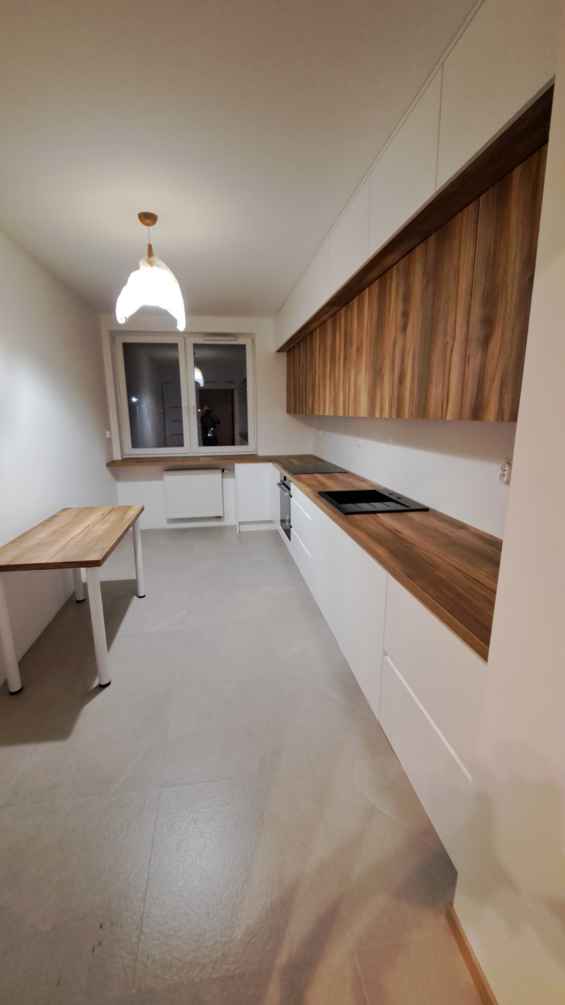 Nowoczesna kuchnia w bieli i drewnie: białe szafki dolne z frezowanymi uchwytami, drewniane blaty i szafki górne, czarna płyta grzewcza i zlew, drewniany stół z białymi nogami, lampa wisząca...