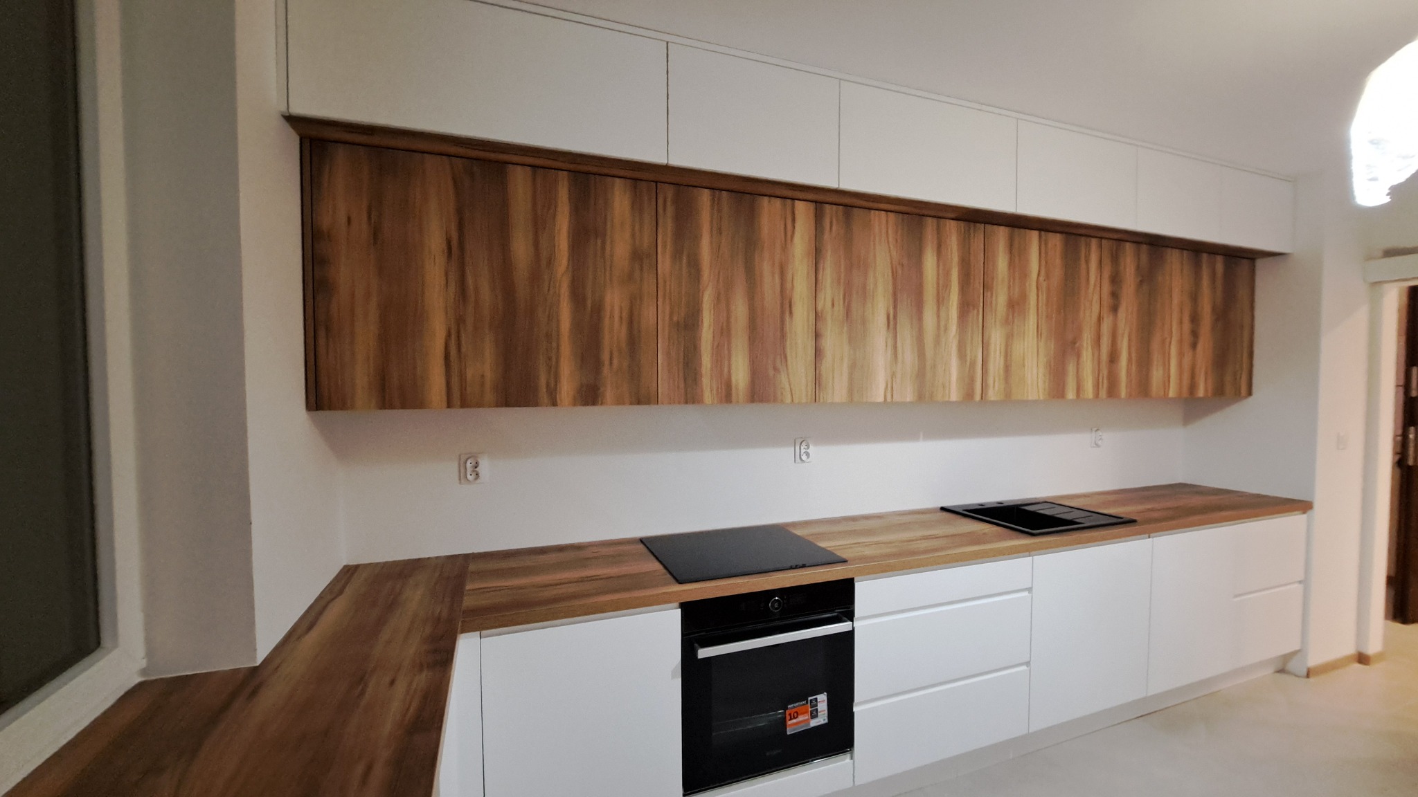 Nowoczesna kuchnia z białymi szafkami dolnymi i drewnianymi szafkami wiszącymi, drewniany blat z płytą indukcyjną i piekarnikiem.