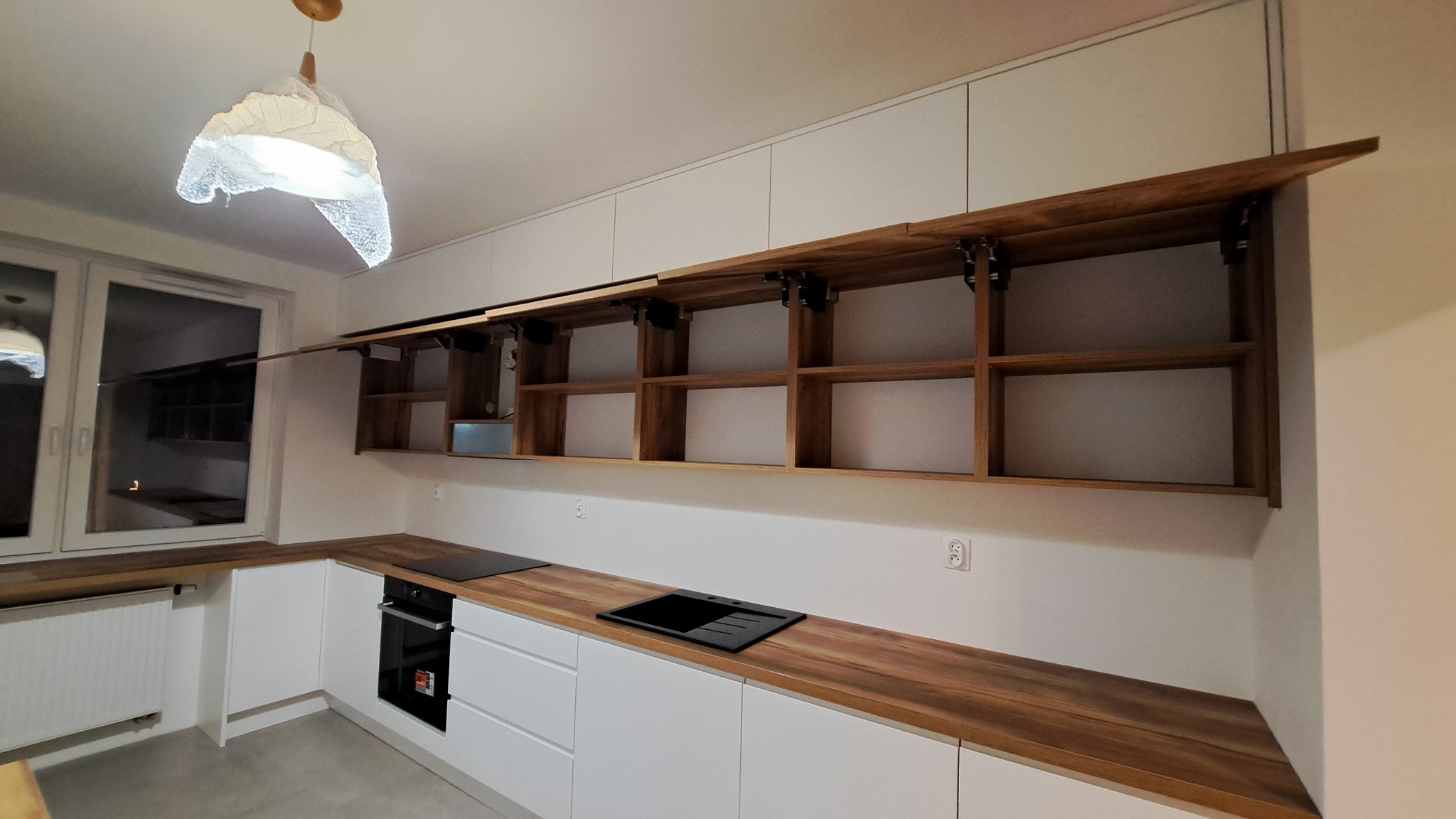 Nowoczesna kuchnia z białymi szafkami dolnymi i drewnianymi blatami oraz górnymi szafkami z drewnianym wnętrzem, otwierane do góry.