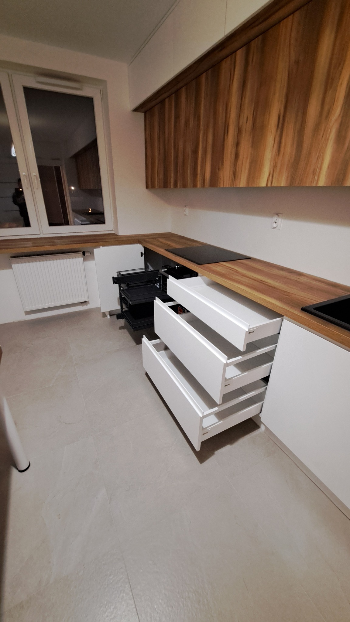 Nowoczesna kuchnia z białymi szafkami i drewnianym blatem, otwarte szuflady z systemem cargo, widoczny fragment okna i grzejnika.