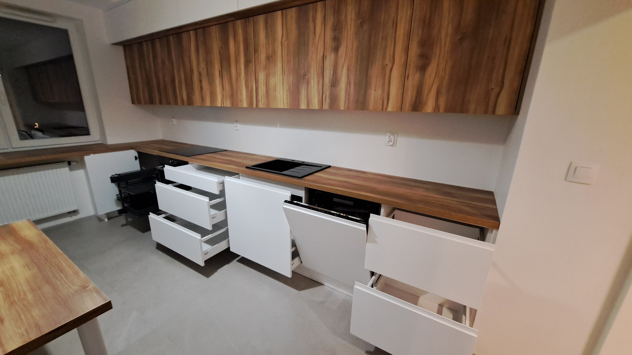 Nowoczesna kuchnia z drewnianym blatem i białymi frontami szafek, niektóre szuflady i szafki otwarte, ukazujące wnętrze.