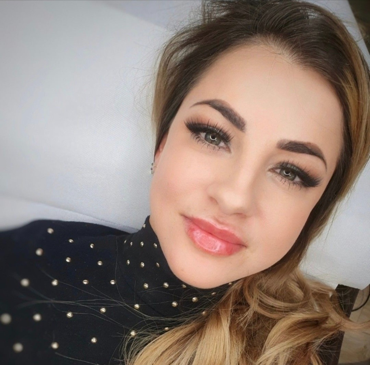 Portret kobiety z wyraźnie podkreślonymi, długimi rzęsami i błyszczącymi ustami, leżącej na białym tle.