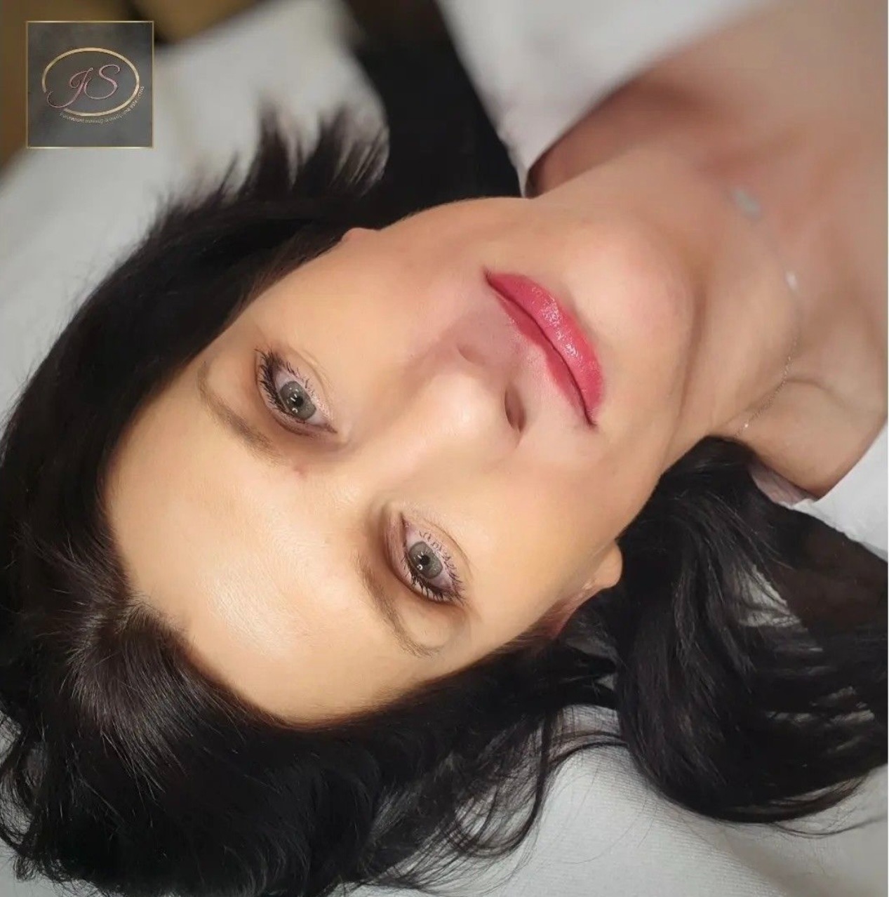 Kobieta leżąca na plecach z wykonanym makijażem permanentnym ust w odcieniu czerwieni, widoczne delikatne podkreślenie konturu i wypełnienie kolorem. W tle logo firmy.