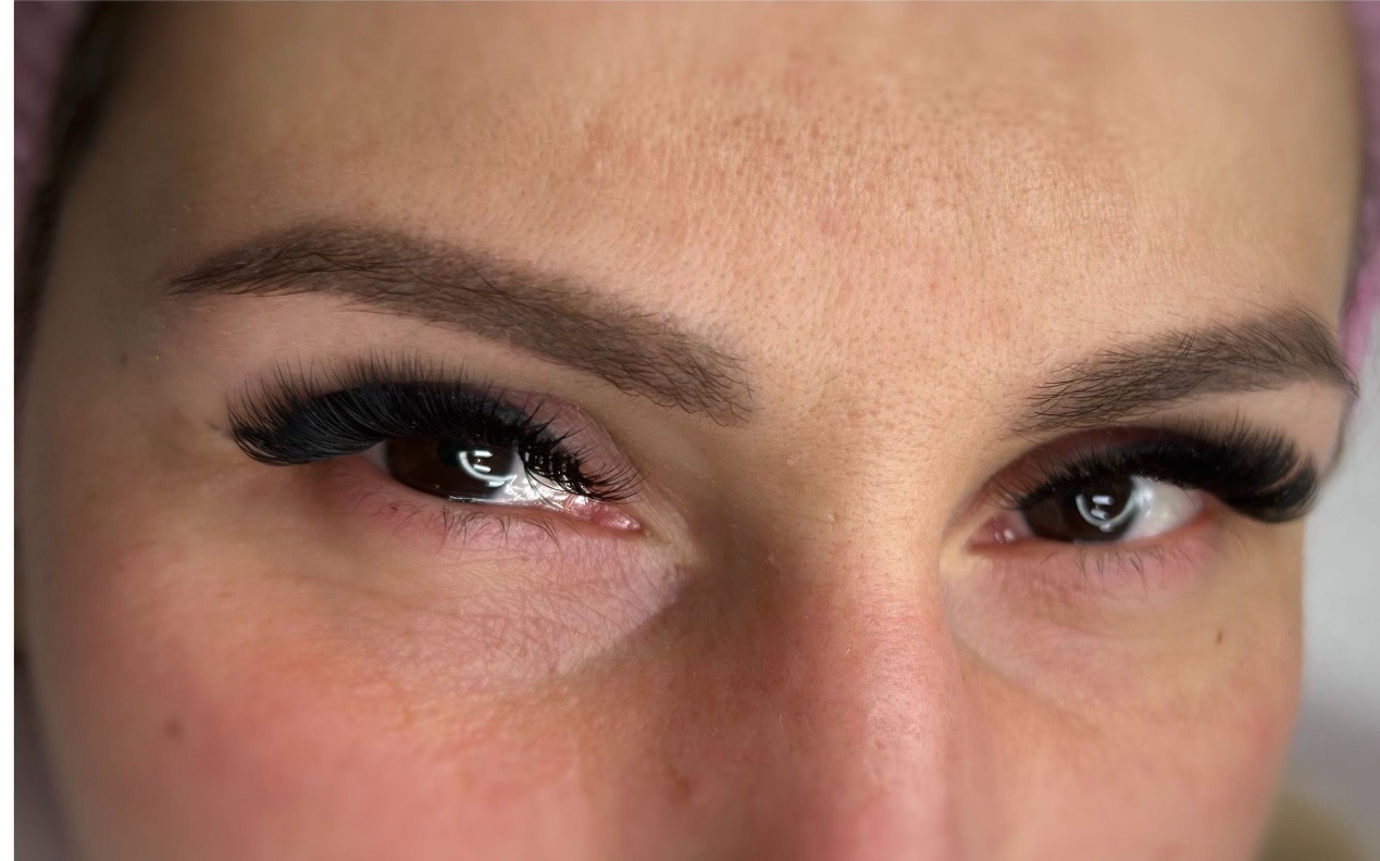 Zbliżenie na twarz kobiety z wyraźnie podkreślonymi, długimi, czarnymi rzęsami, brwi starannie wyregulowane, skóra o naturalnej fakturze, widoczne odbicie światła w źrenicach.